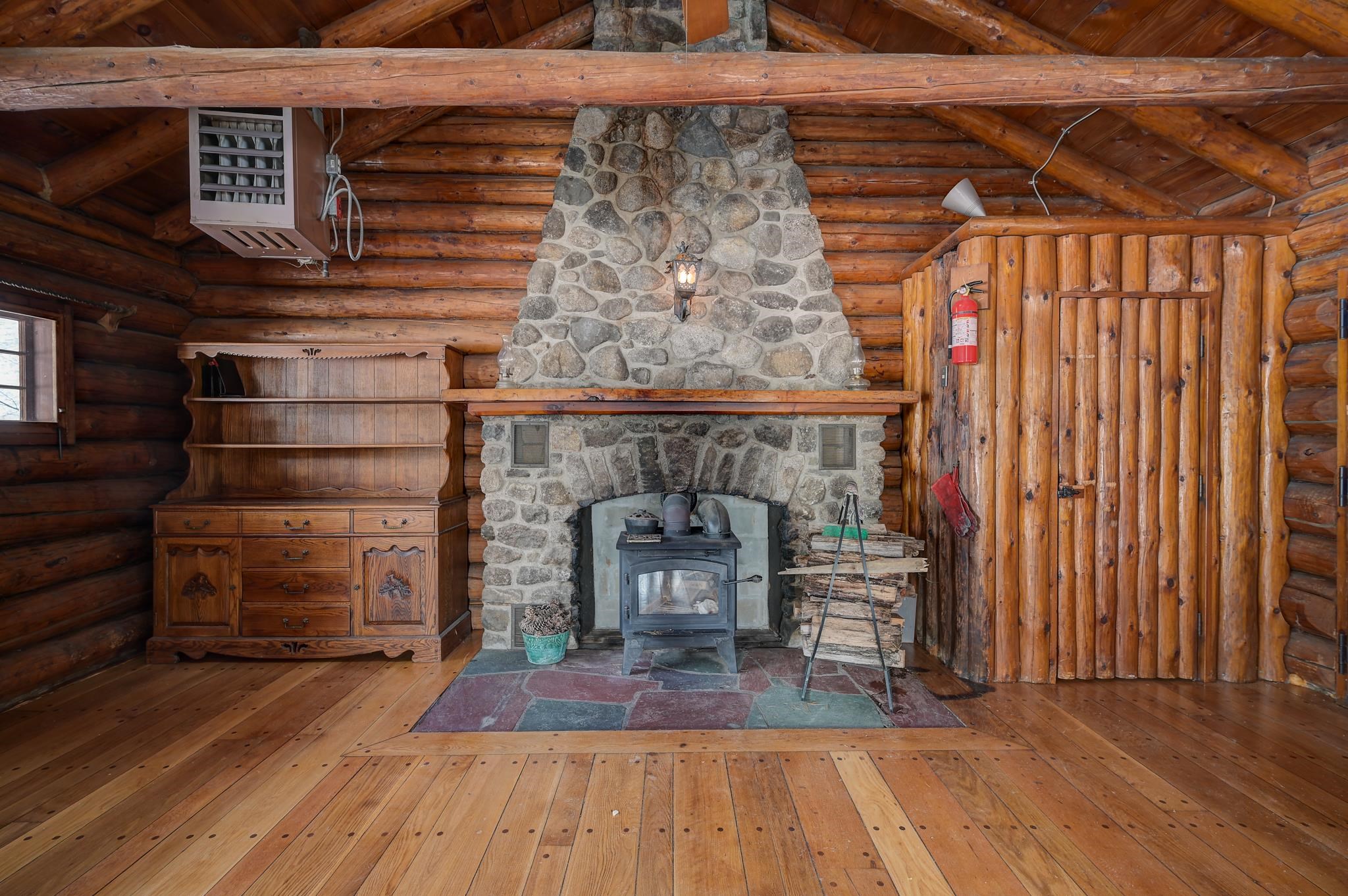 Log cabin living area with a tall stone fireplace, wood-burning stove, and rustic plank door.
