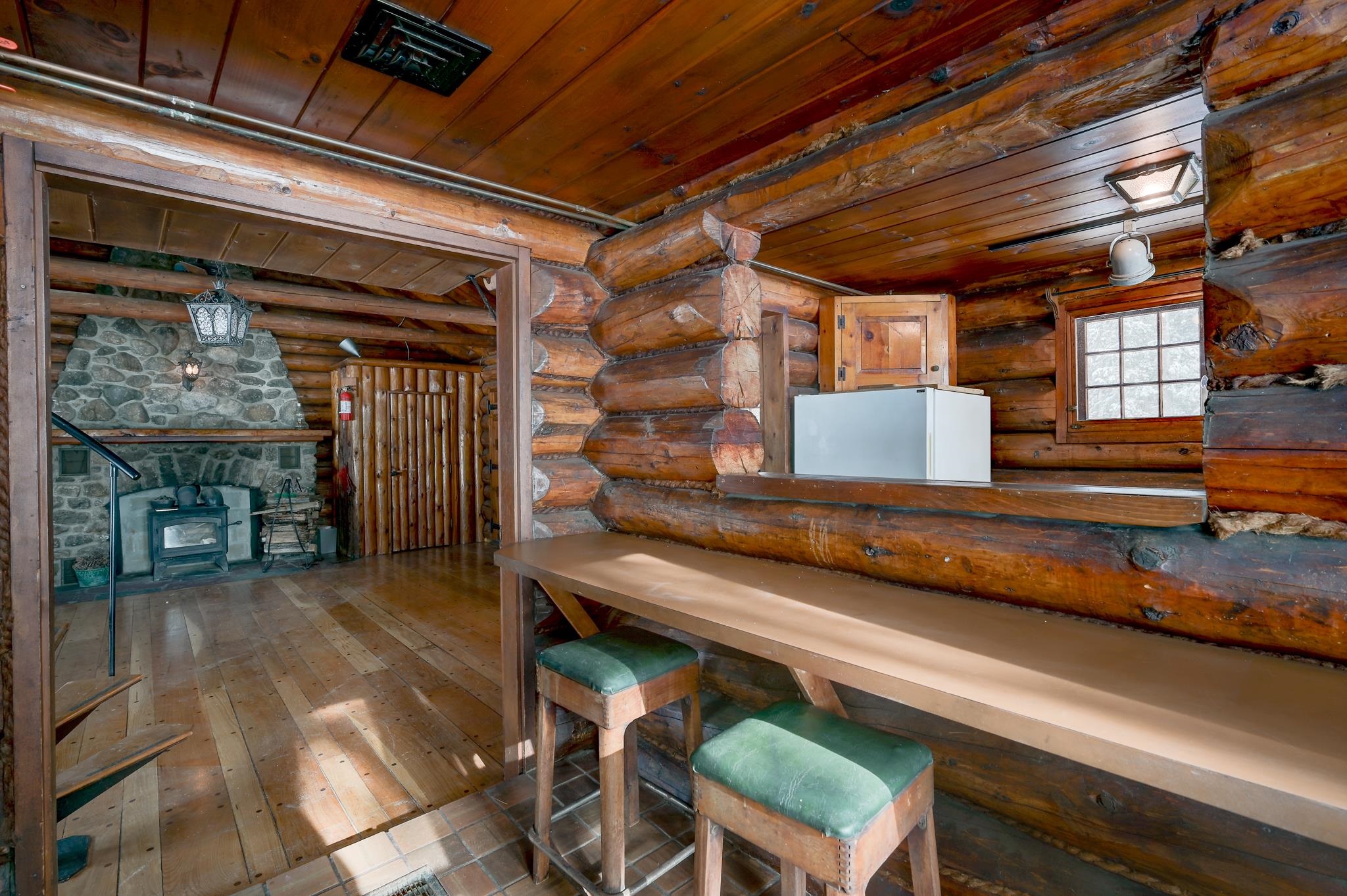Rustic log cabin interior with a stone fireplace, wood stove, and long counter with stools.