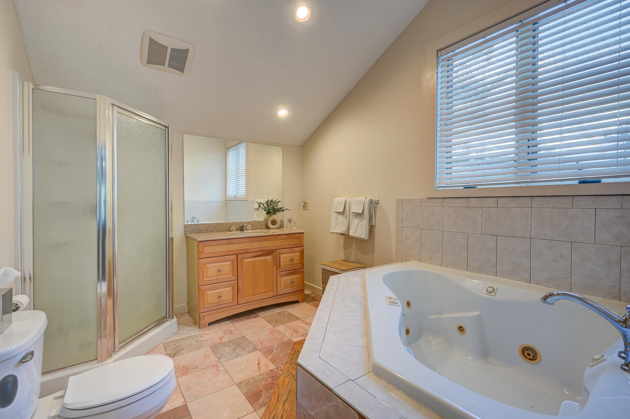Bathroom with a jetted soaking tub beneath a wide window, glass shower, and wood vanity.