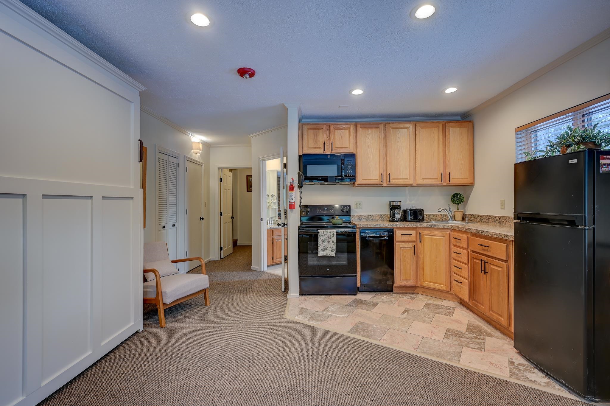 Condo kitchen with wood cabinets and black appliances, opening onto a carpeted sitting area.