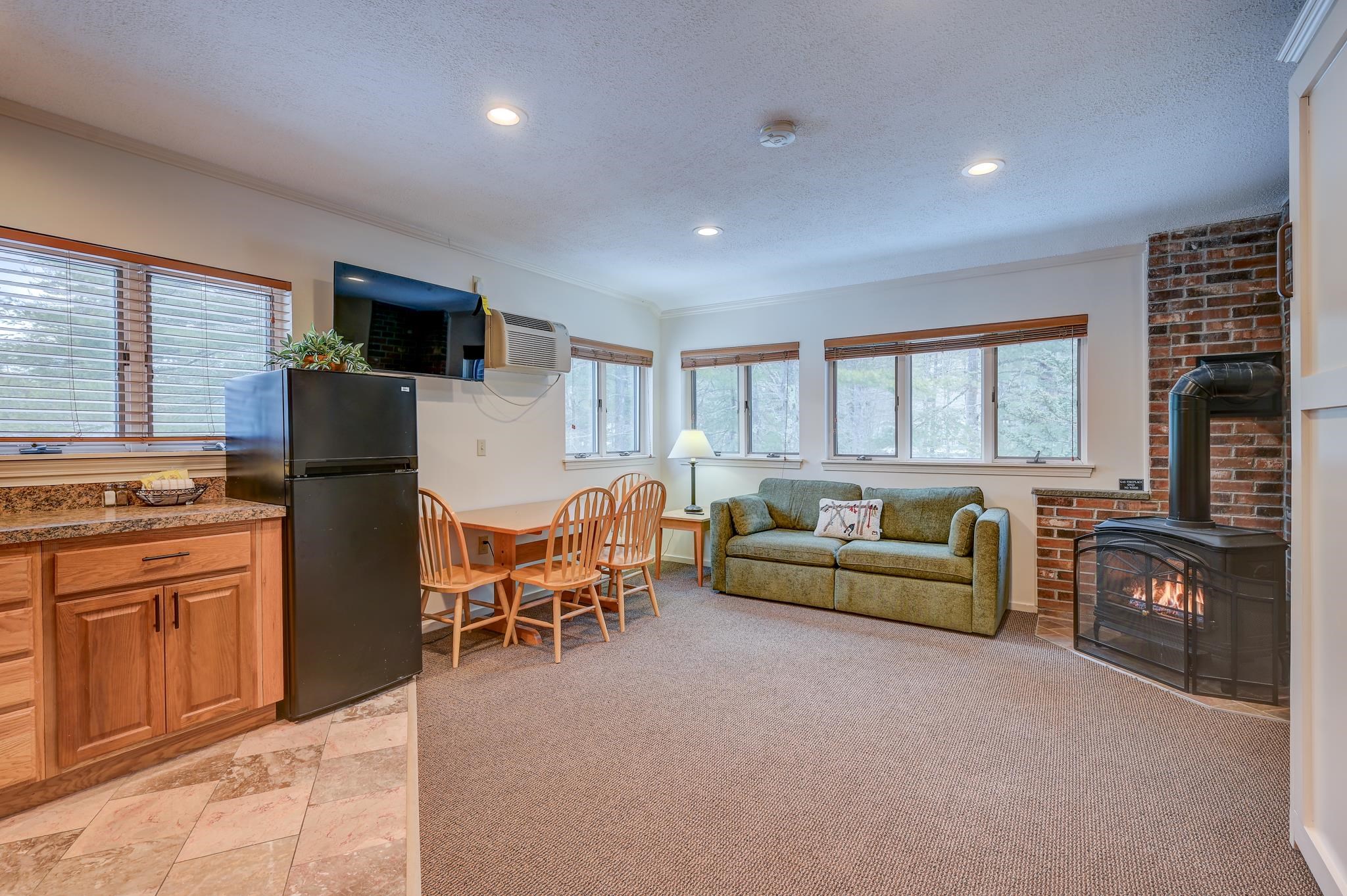 Furnished living and dining area with a green sofa and lit wood stove beside a compact kitchen.