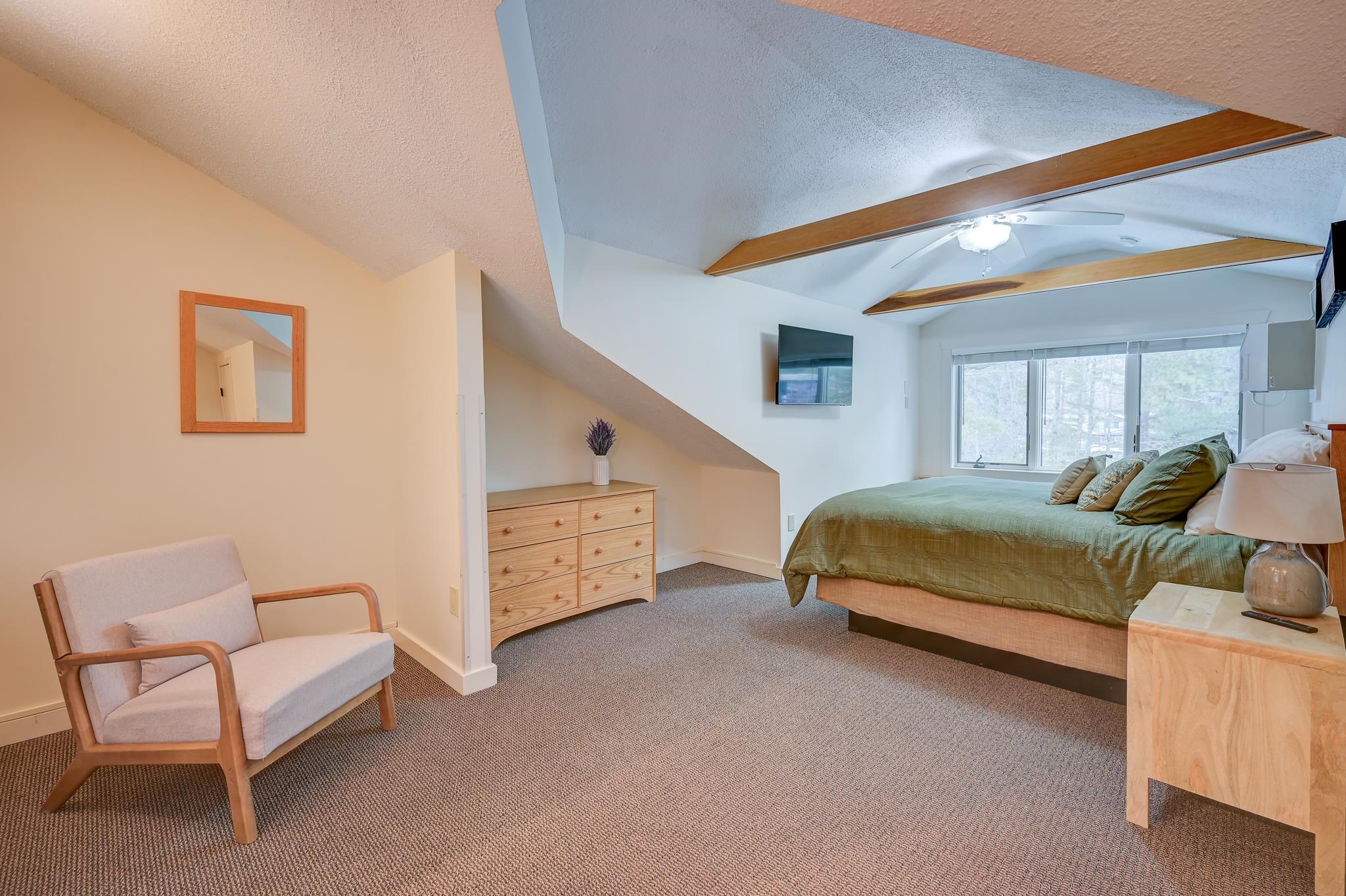 Bedroom with vaulted wood-beamed ceiling, green bedspread, wide windows, dresser, and upholstered chair.