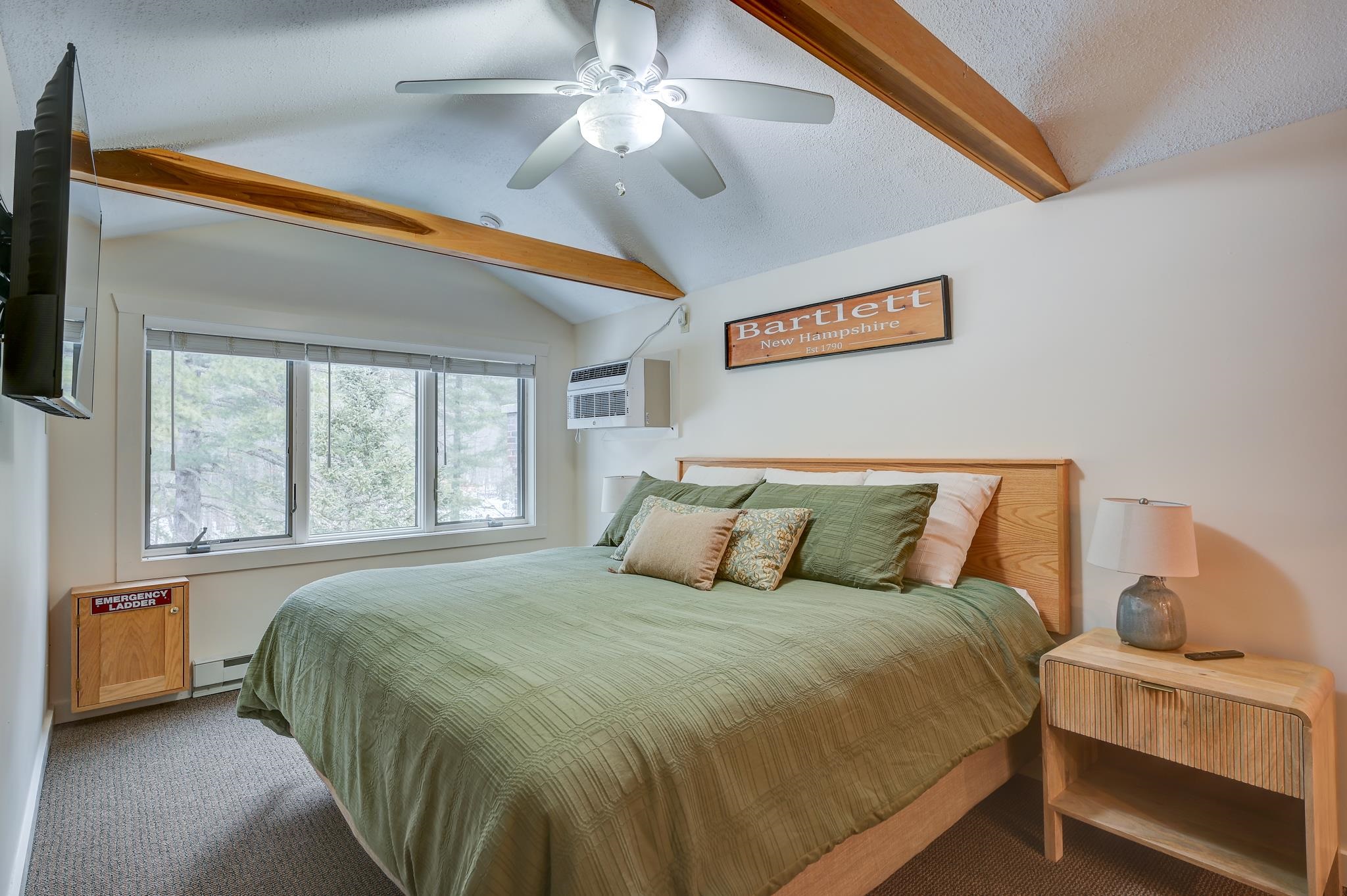 Furnished bedroom with a green-covered king bed, vaulted wood-beam ceiling, and large window overlooking trees.