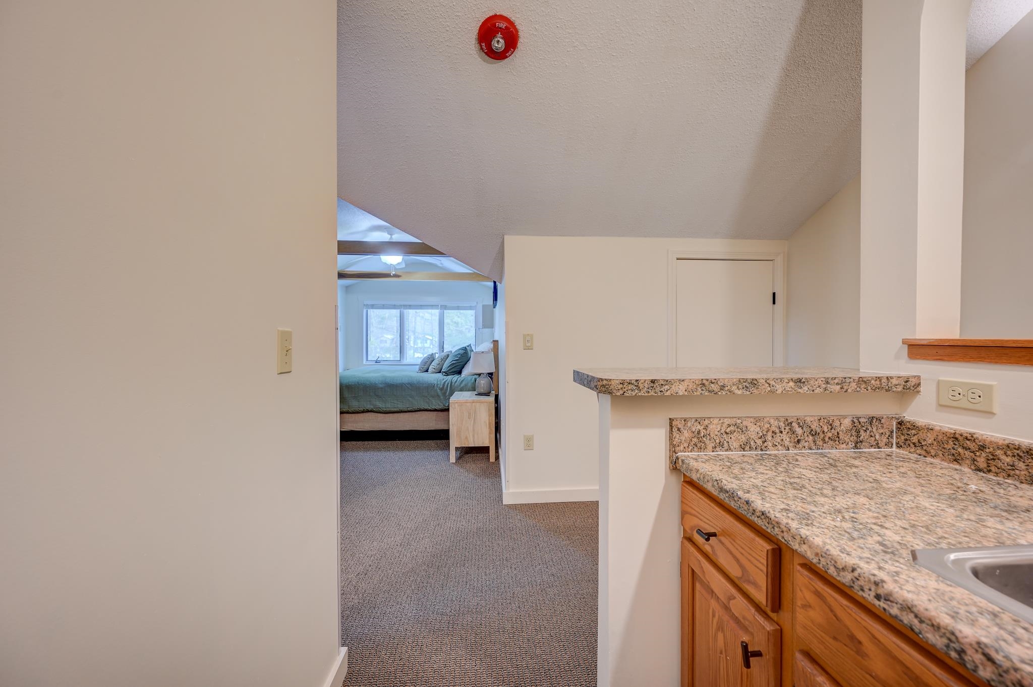 Granite kitchen counter and wood cabinets beside a hallway leading to a bedroom with a green bedspread.