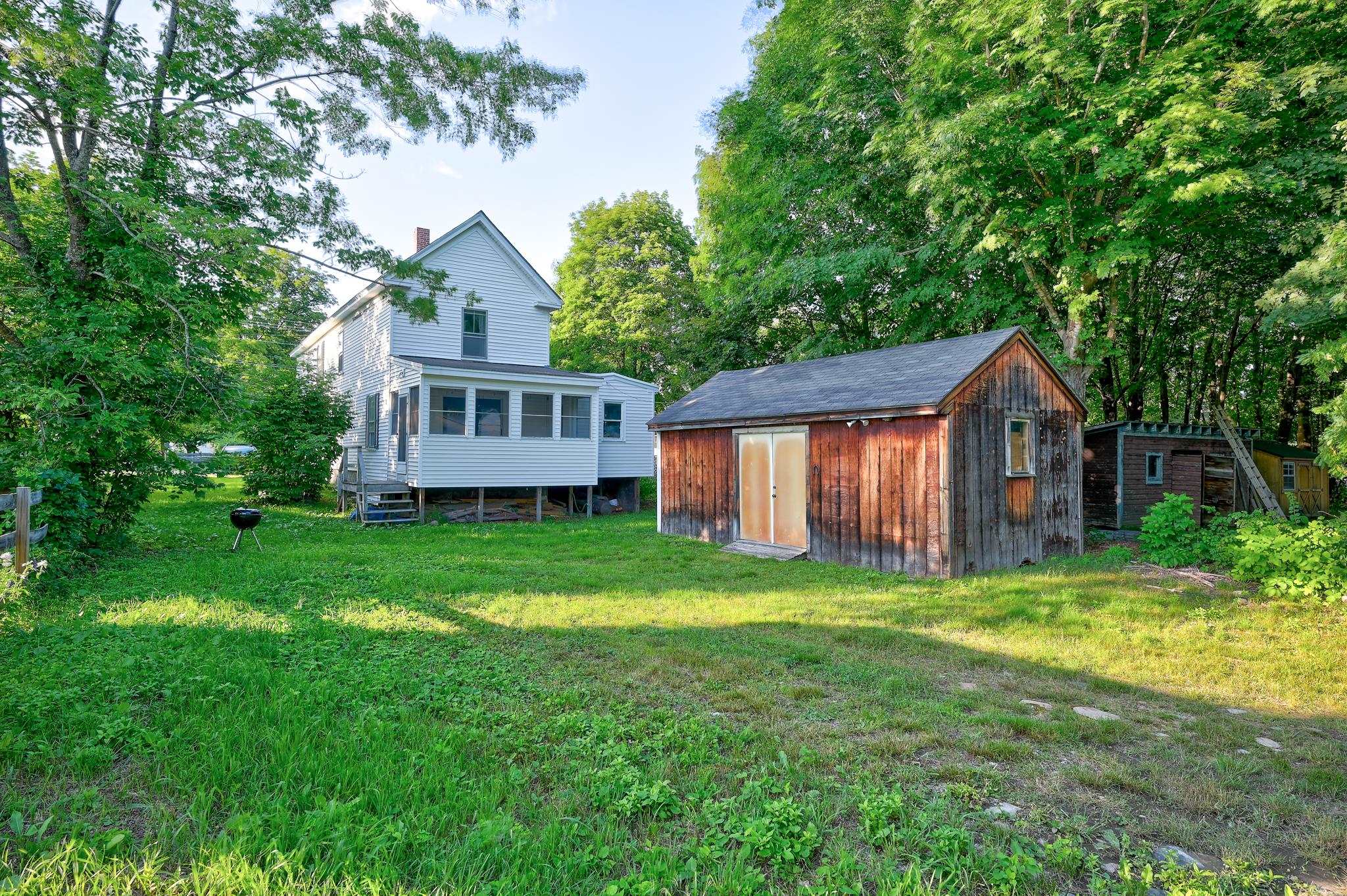 White two-story house with an enclosed porch and wooden shed in a tree-lined yard.