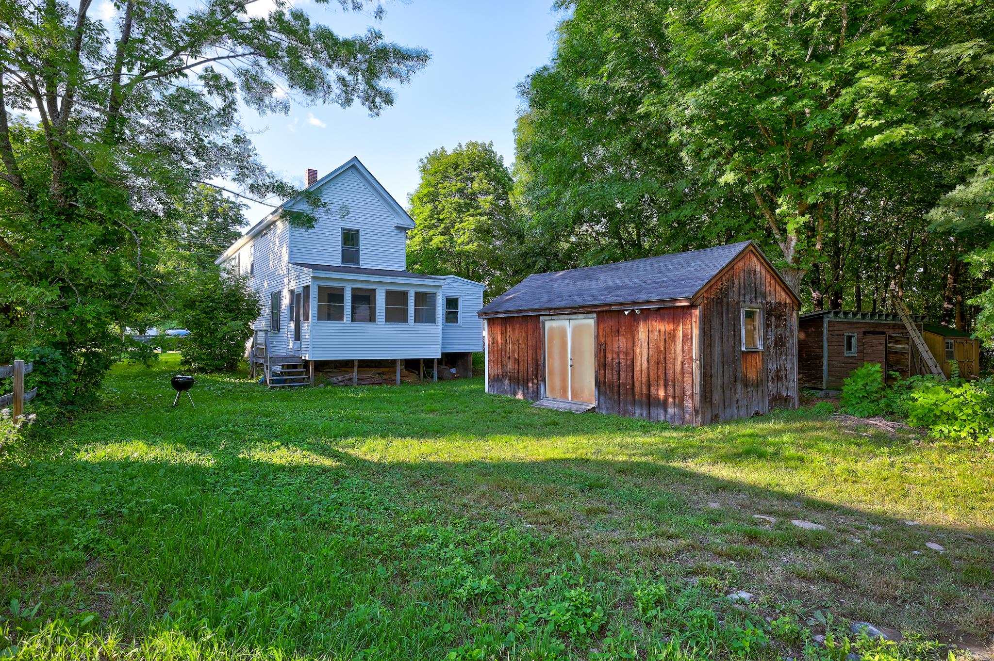 White house with an enclosed porch beside a large wooden shed in a leafy backyard.