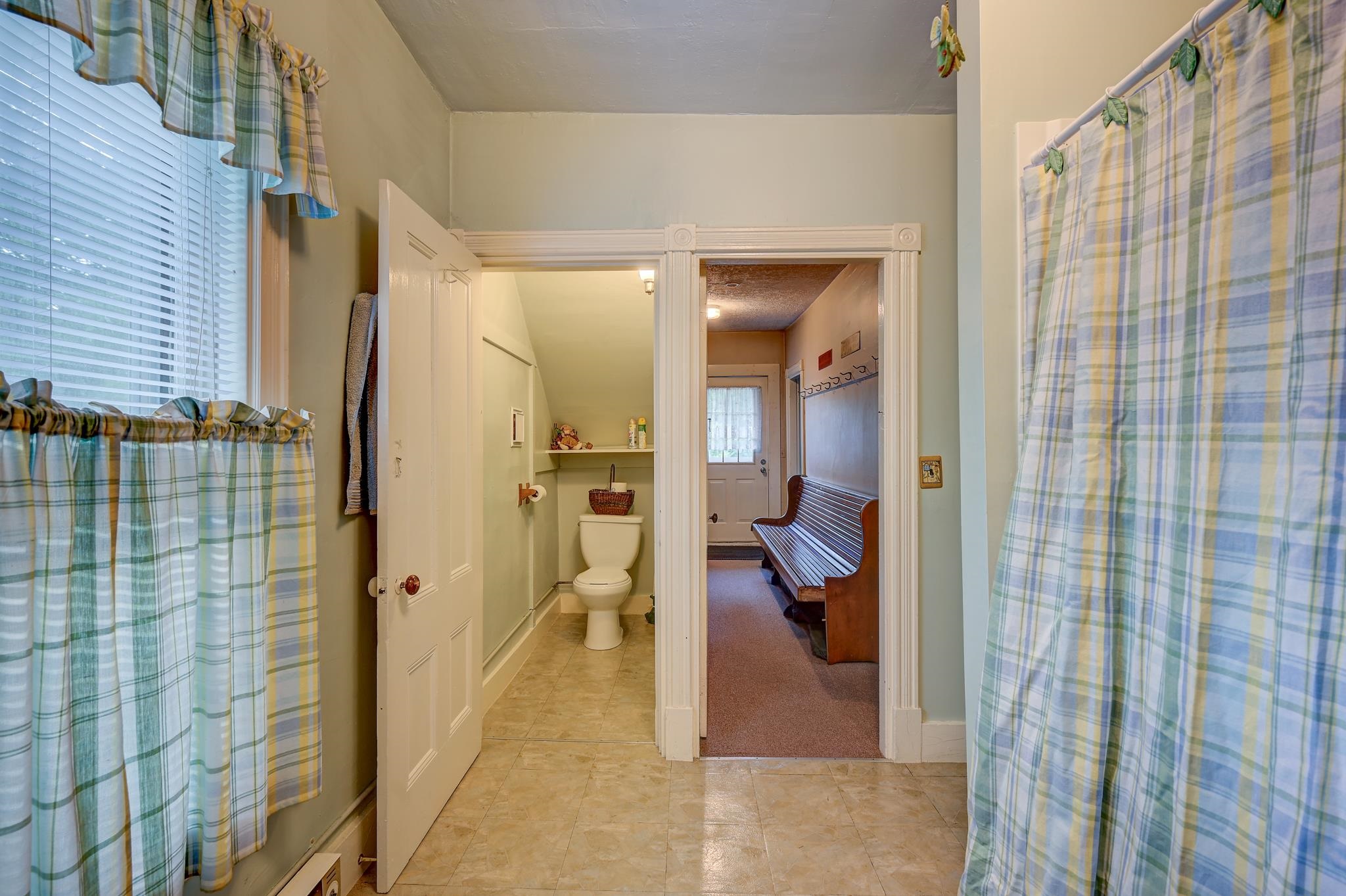 Bathroom with plaid shower curtain, toilet alcove, and doorway to a bench-lined hallway.