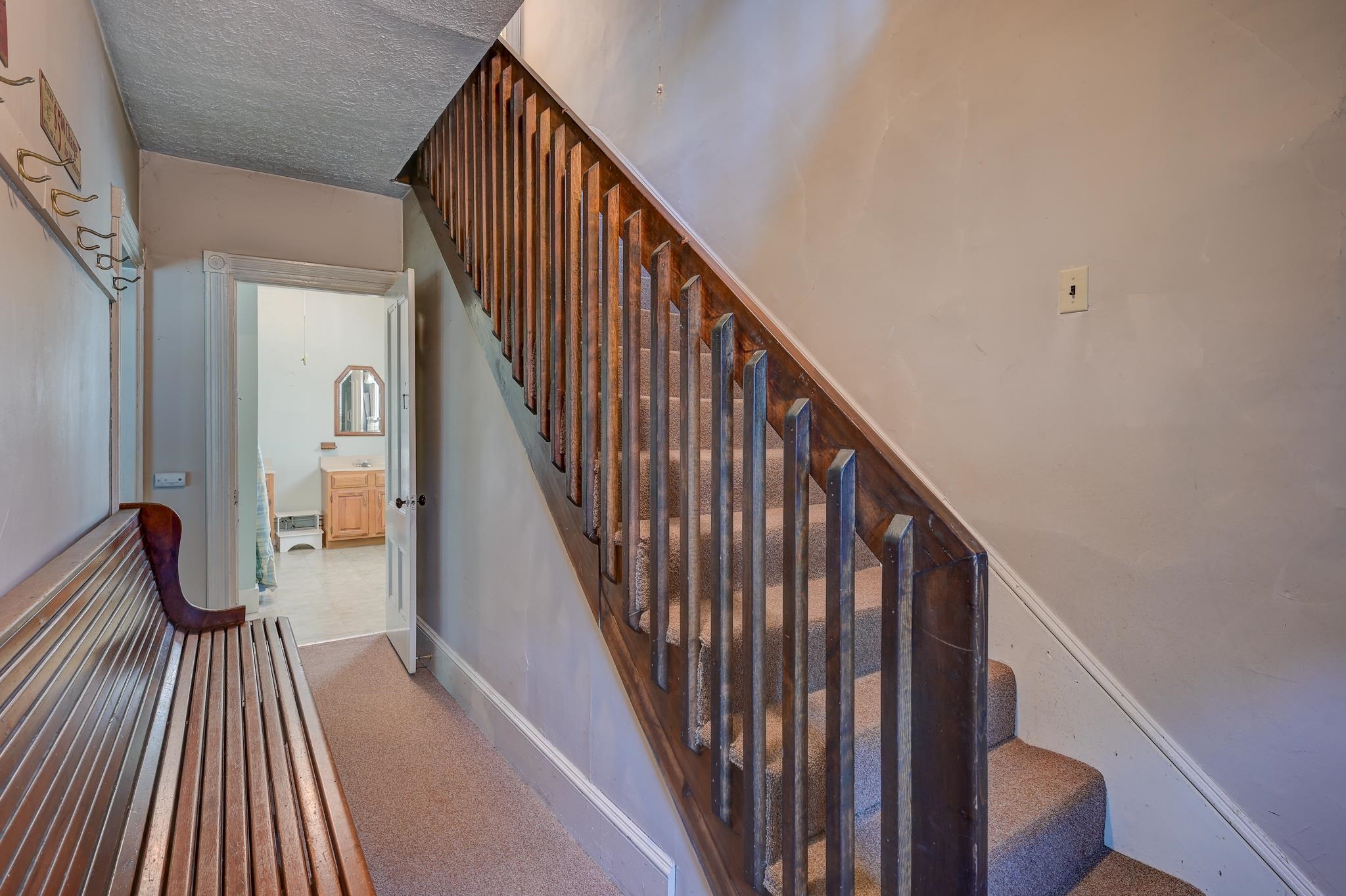 Hallway with a carpeted staircase, dark wood railing, and long wooden bench beside an open bathroom doorway.
