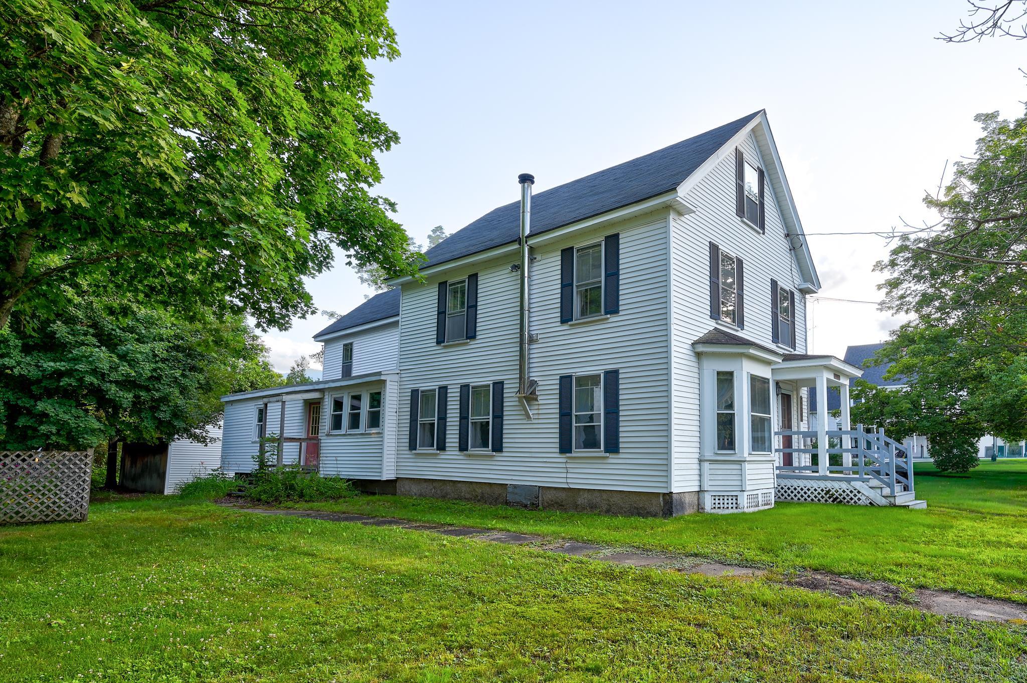 White two-story house with dark shutters, a covered porch, and mature trees around the lawn.