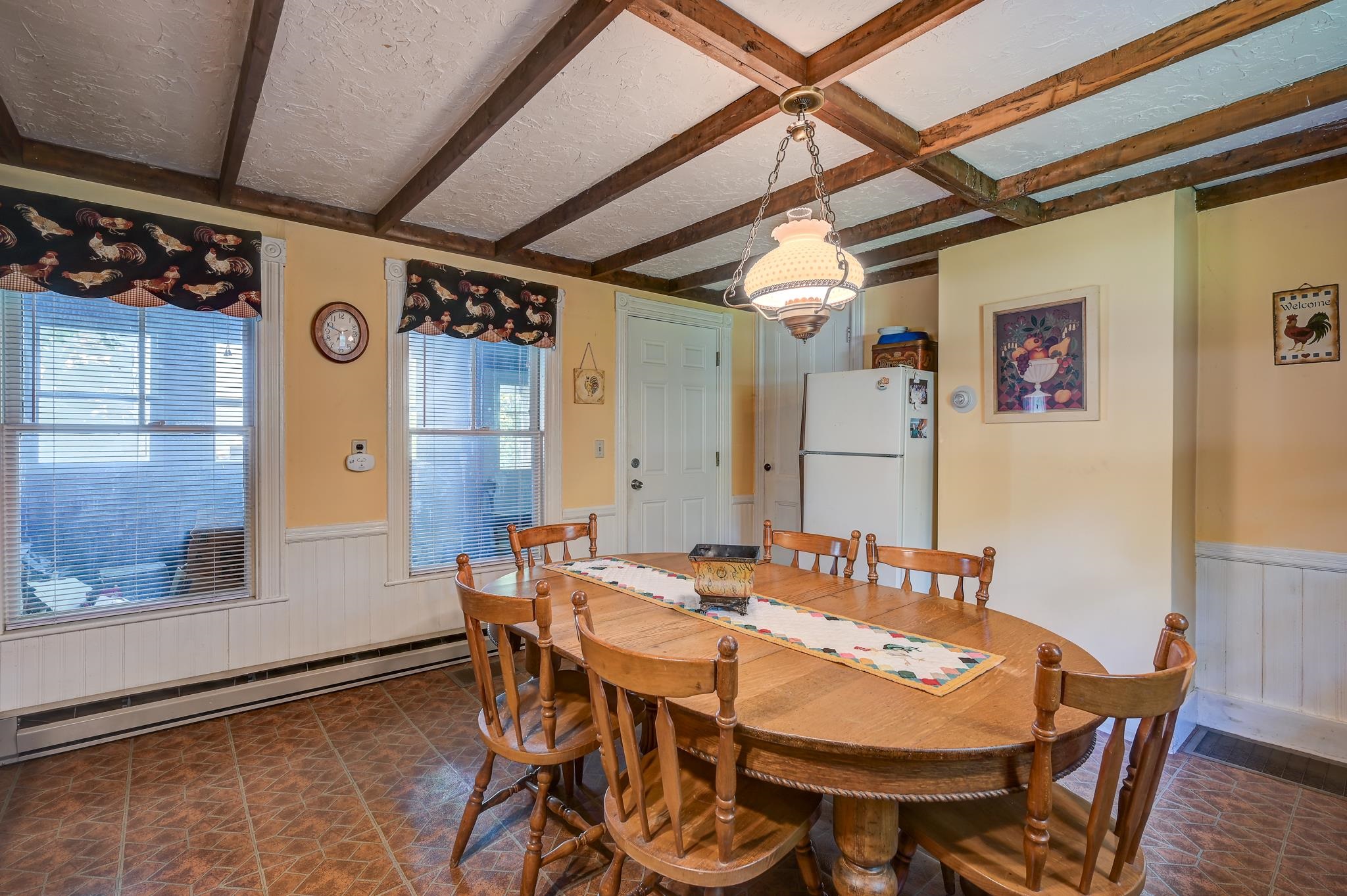 Dining room with an oval wooden table, exposed ceiling beams, and rooster-print window valances.