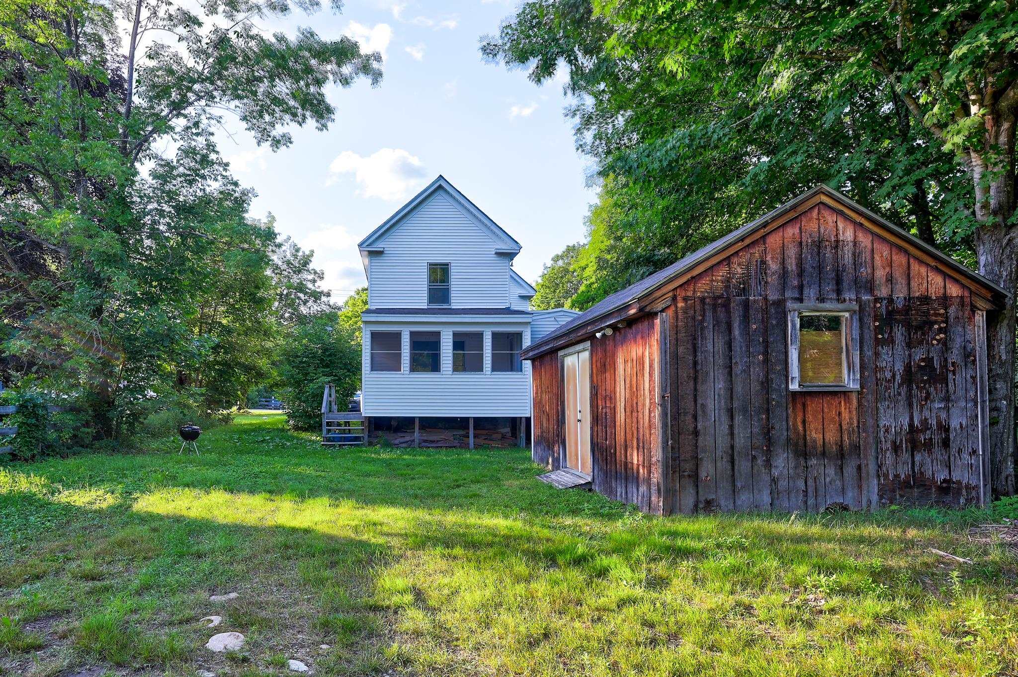 Rear of a white two-story house beside a weathered wooden shed in a leafy backyard.