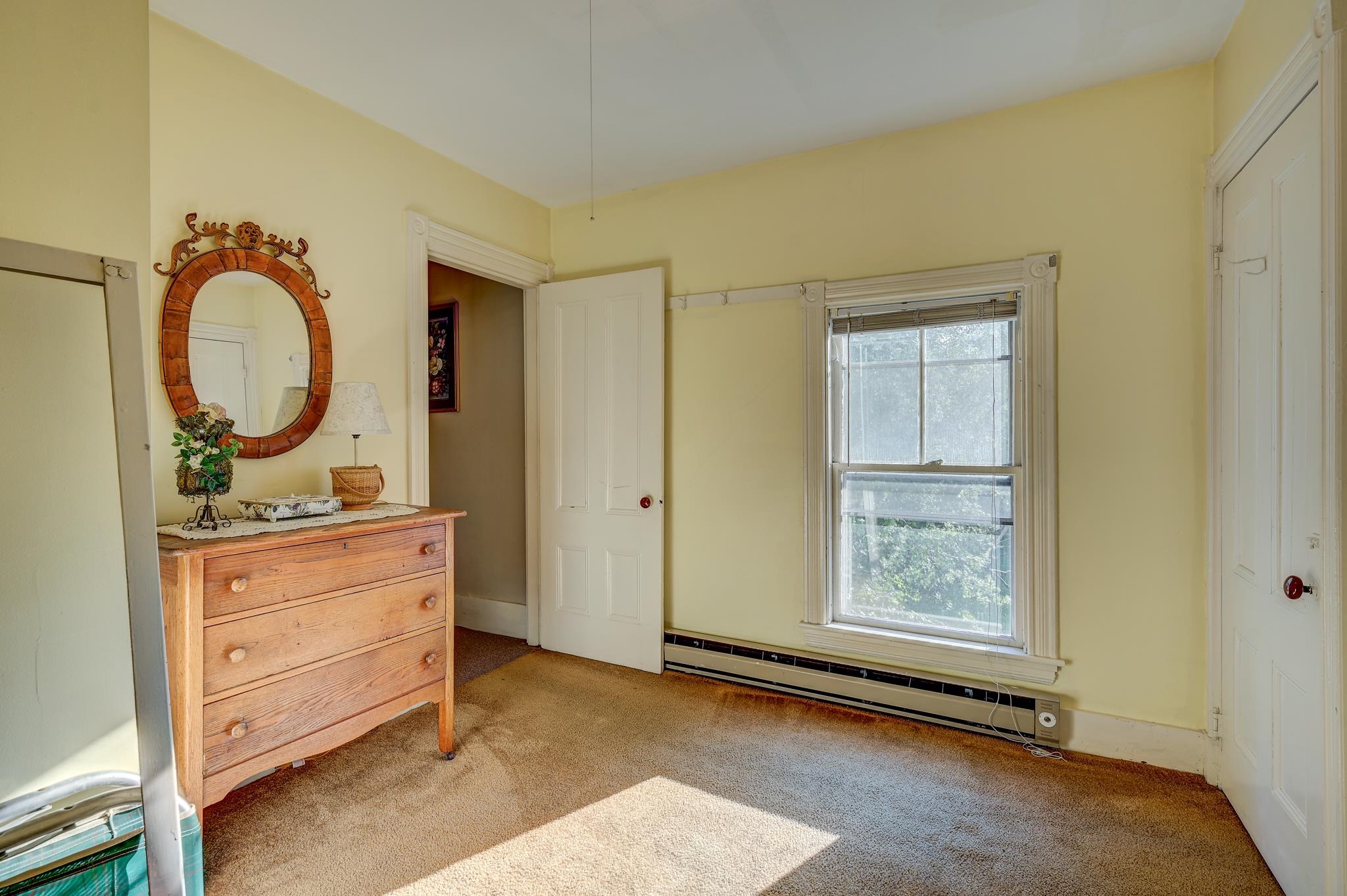 Bedroom with a wooden dresser and oval mirror beside a sunlit window.