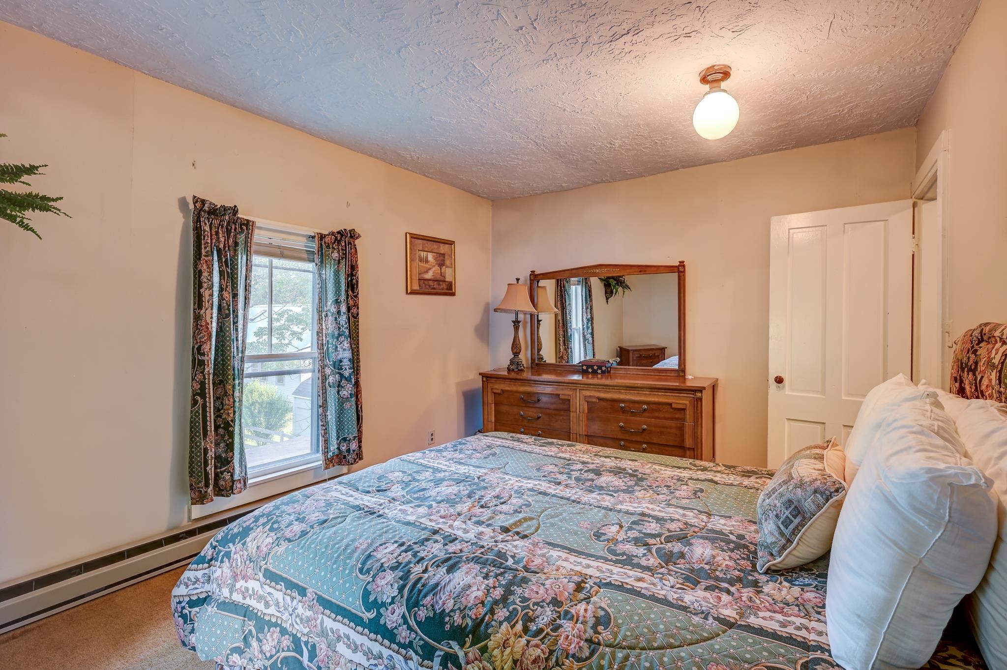 Furnished bedroom with a blue floral quilt, wooden dresser and mirror, and patterned curtains.