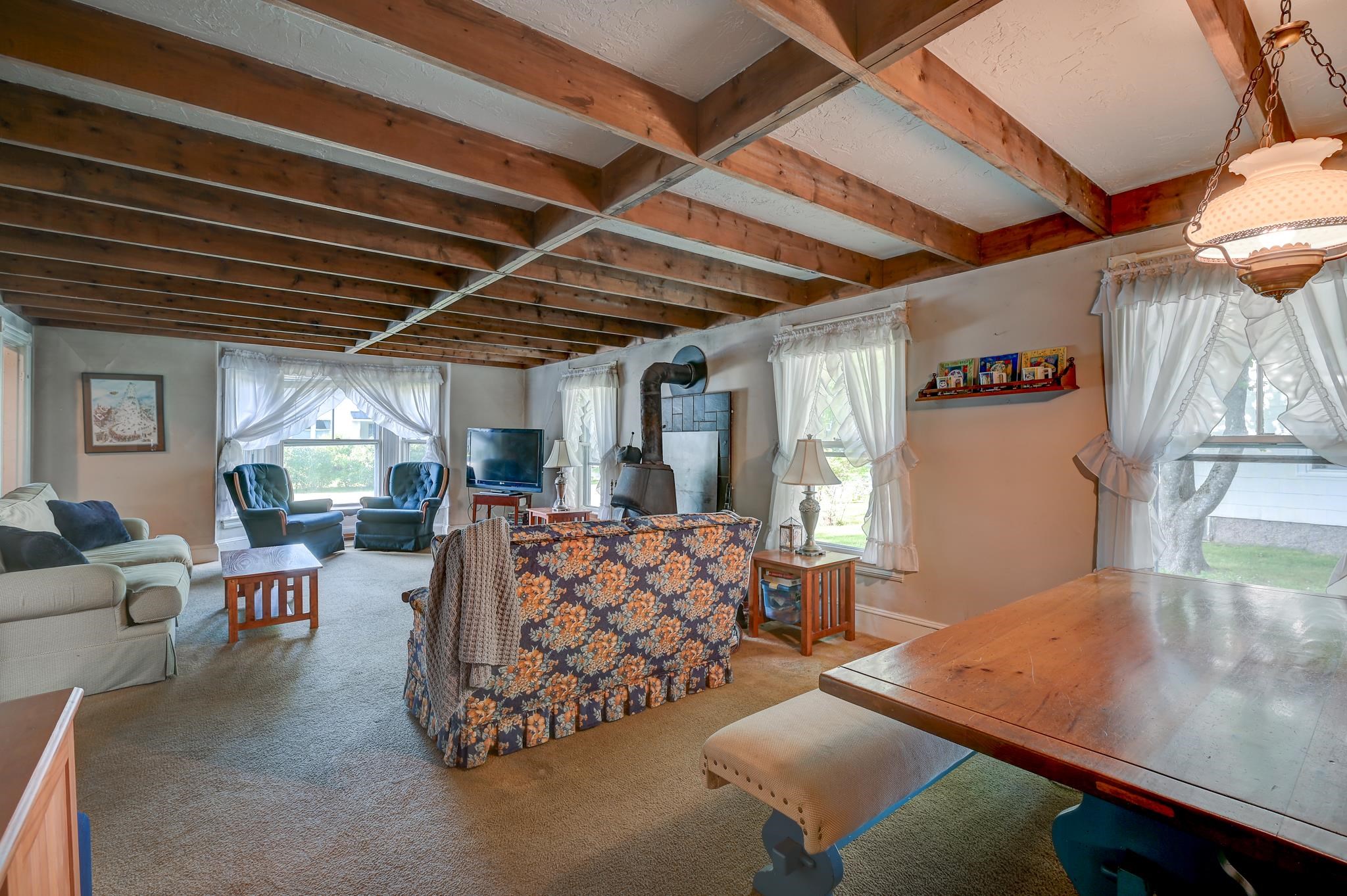 Furnished living room with exposed ceiling beams, a wood stove, and large curtained windows.