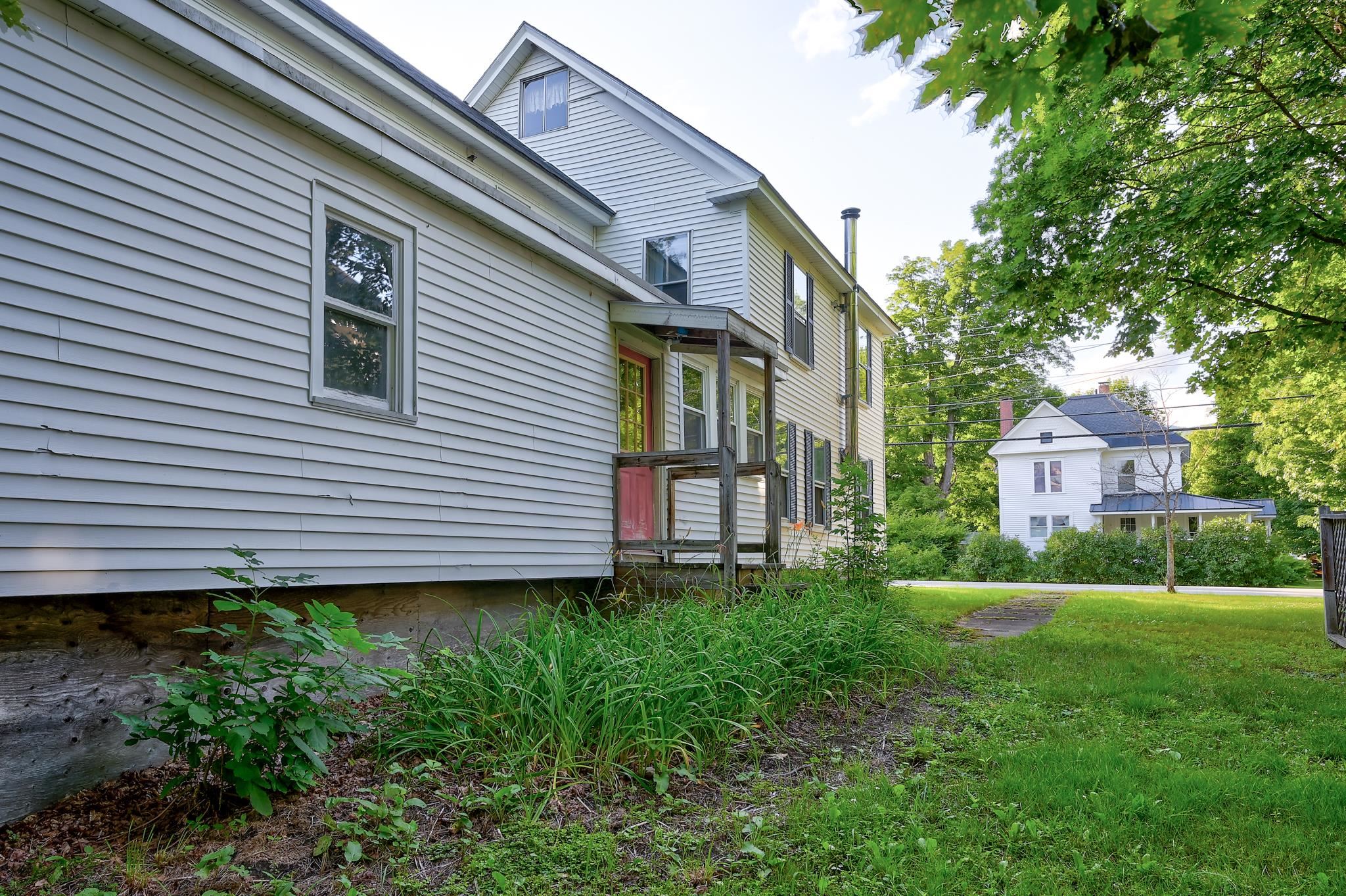 Light-sided multi-story house with a small porch beside a grassy lawn and mature trees.
