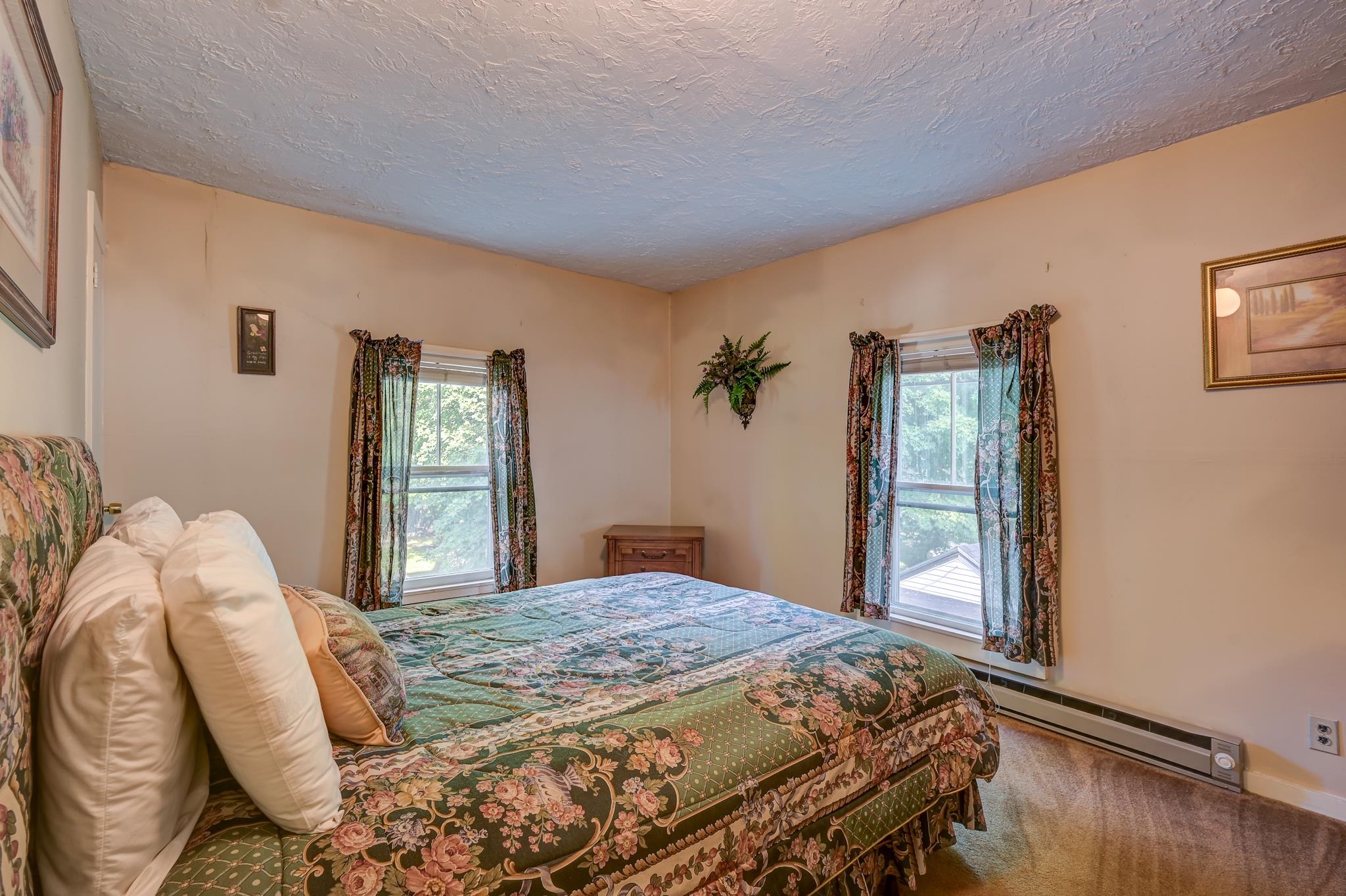 Bedroom with green floral bedding, patterned curtains, and two windows overlooking leafy trees.