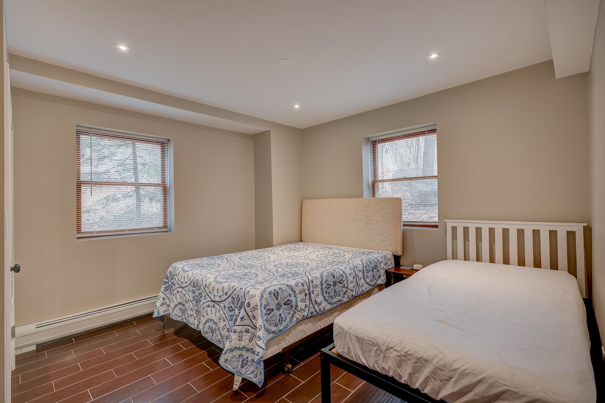 Bedroom with two beds, patterned bedding, wood-look tile floors, and windows with blinds.