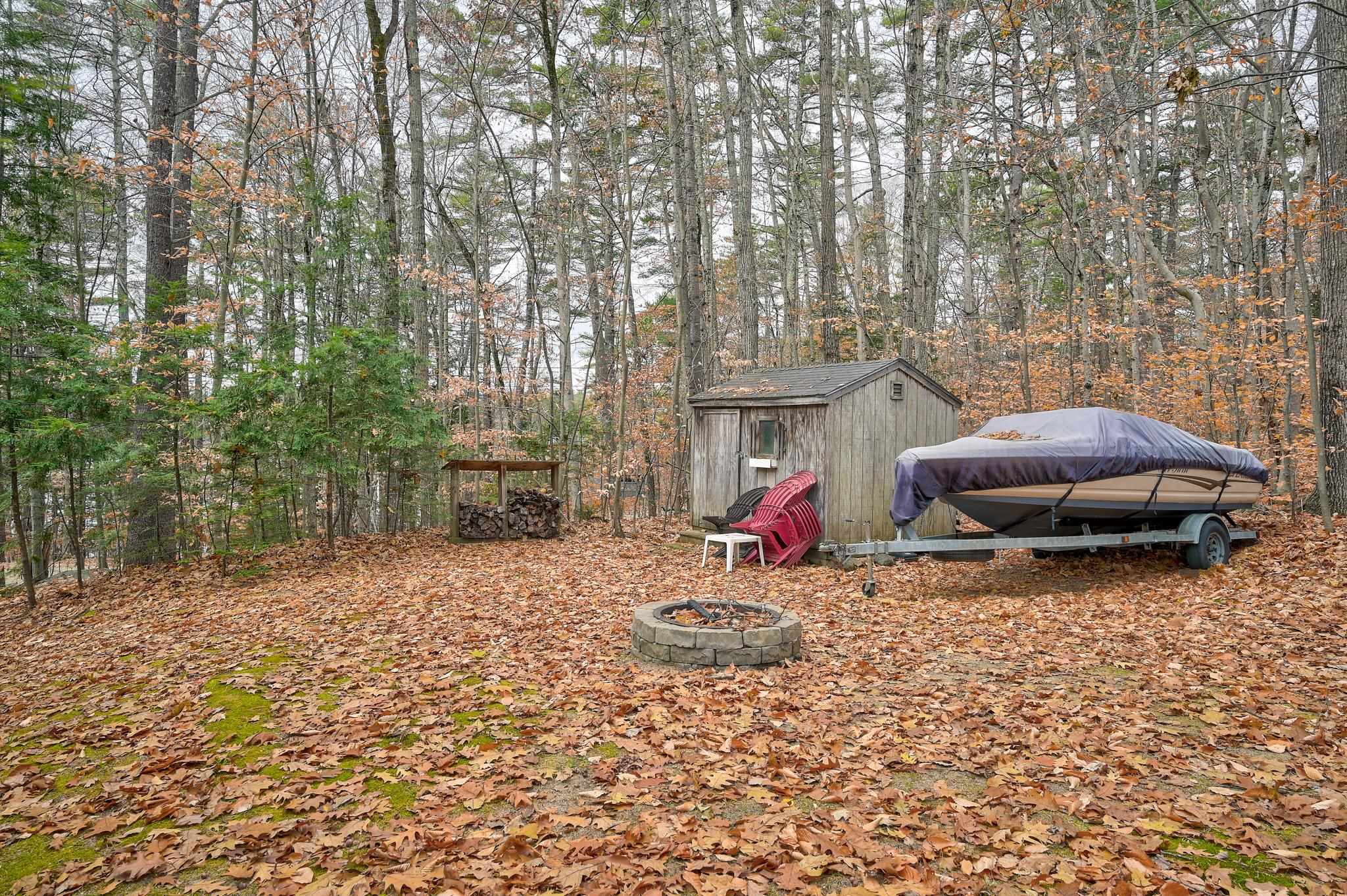 Wooded yard with a gray shed, covered boat, red chairs, and stone fire pit.