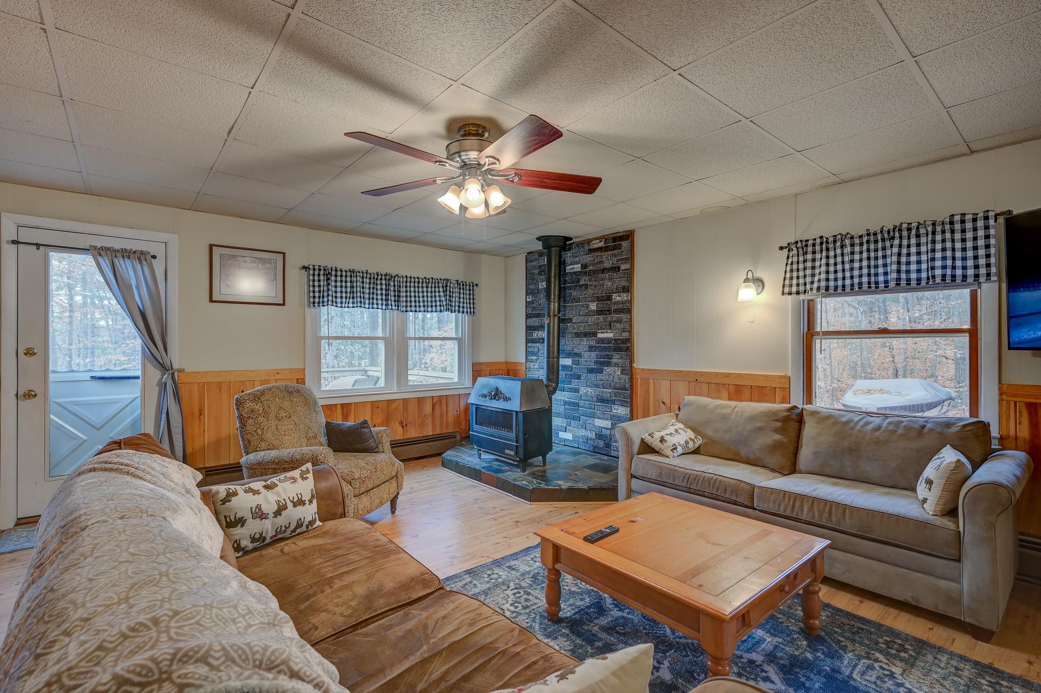 Living room with a brick-surround wood stove, two sofas, and windows overlooking autumn trees.