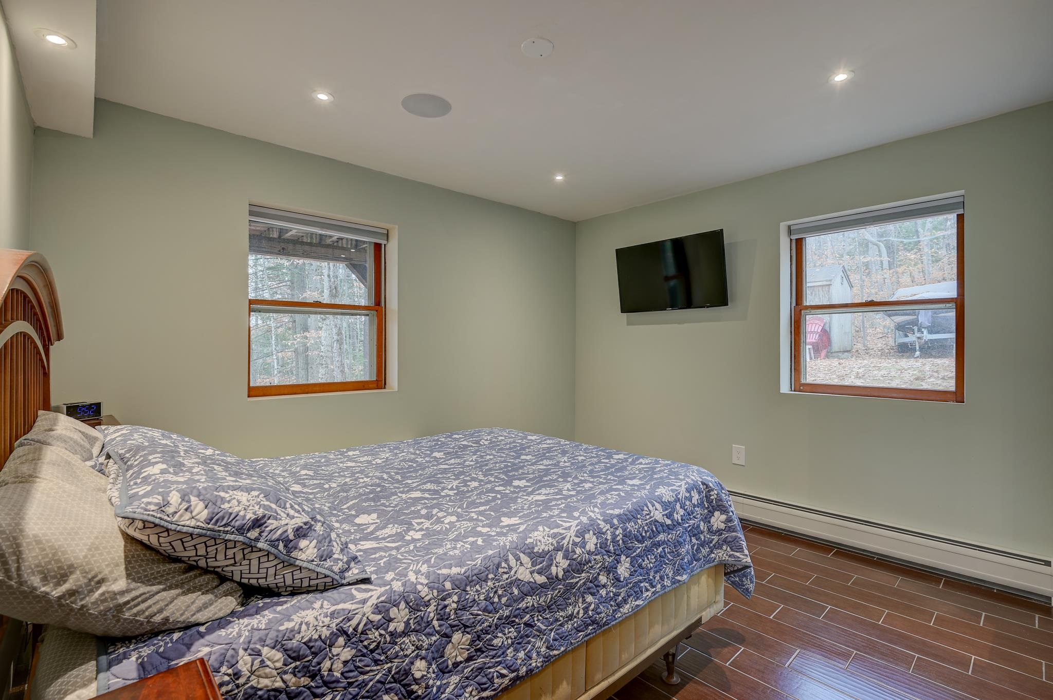 Furnished bedroom with a floral quilt, wall-mounted TV, and two windows overlooking the woods.