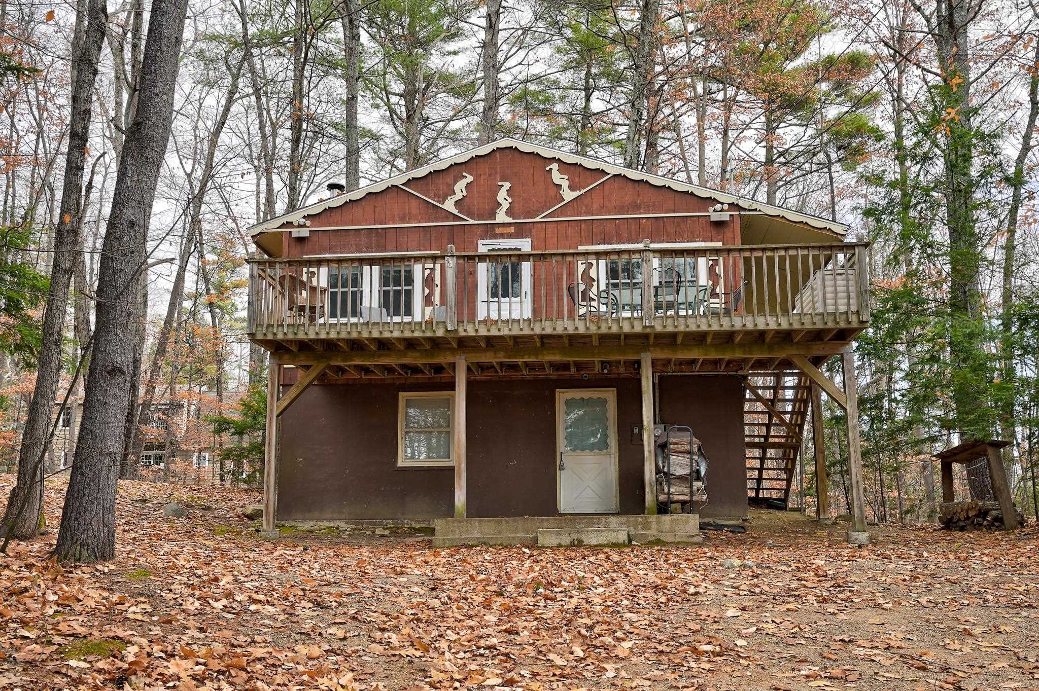 Raised brown cabin with a wide elevated deck and exterior stairs in a leaf-covered woodland.