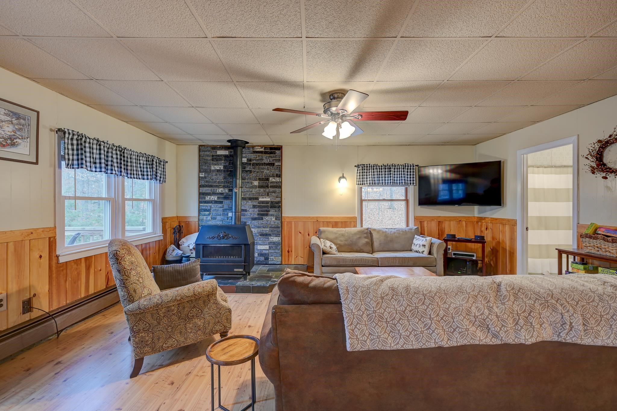 Living room with a black wood stove, wood-paneled walls, large windows, and patterned seating.