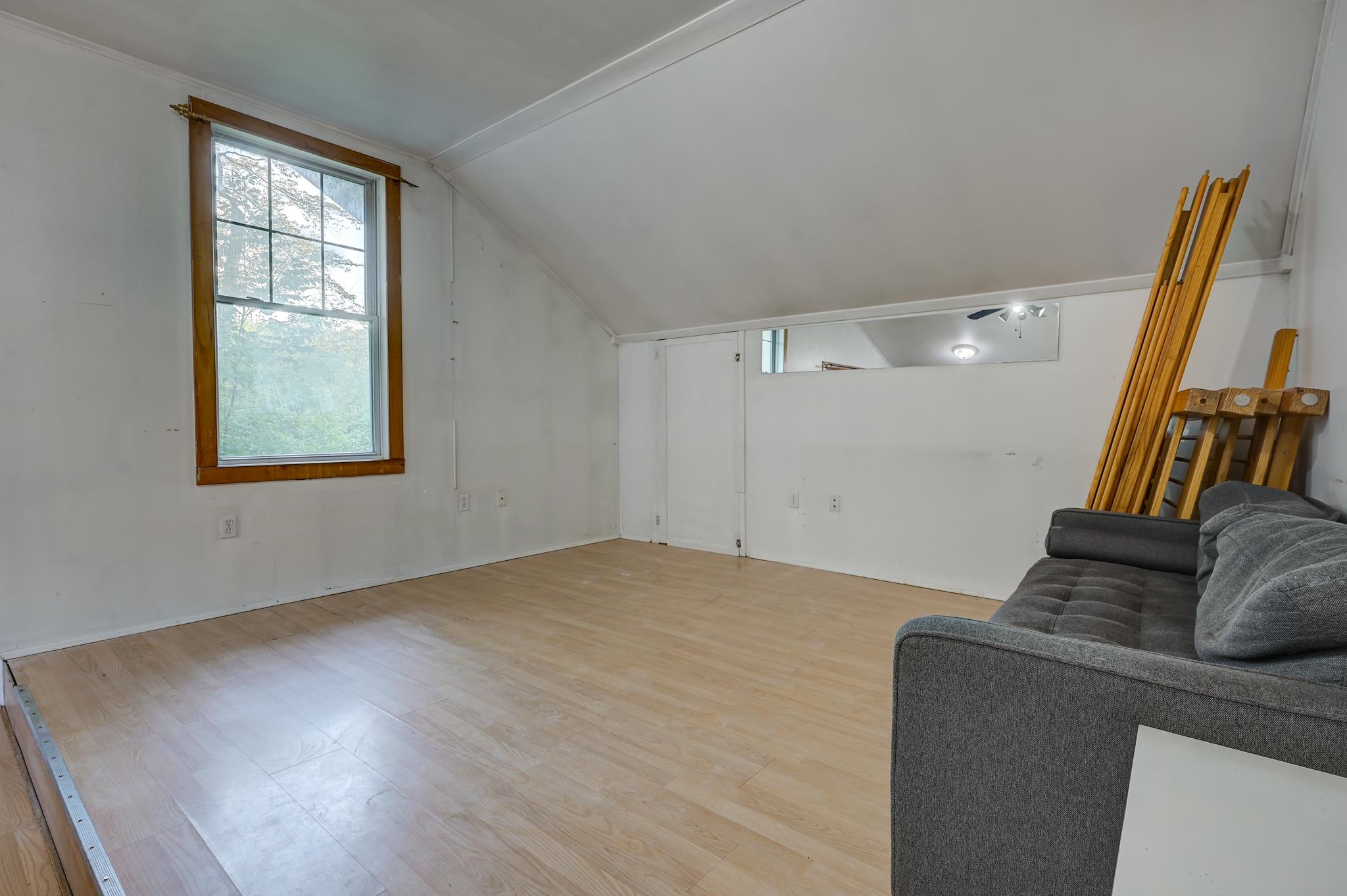 Room with a sloped ceiling, light wood floor, wood-trimmed window, and gray sofa.