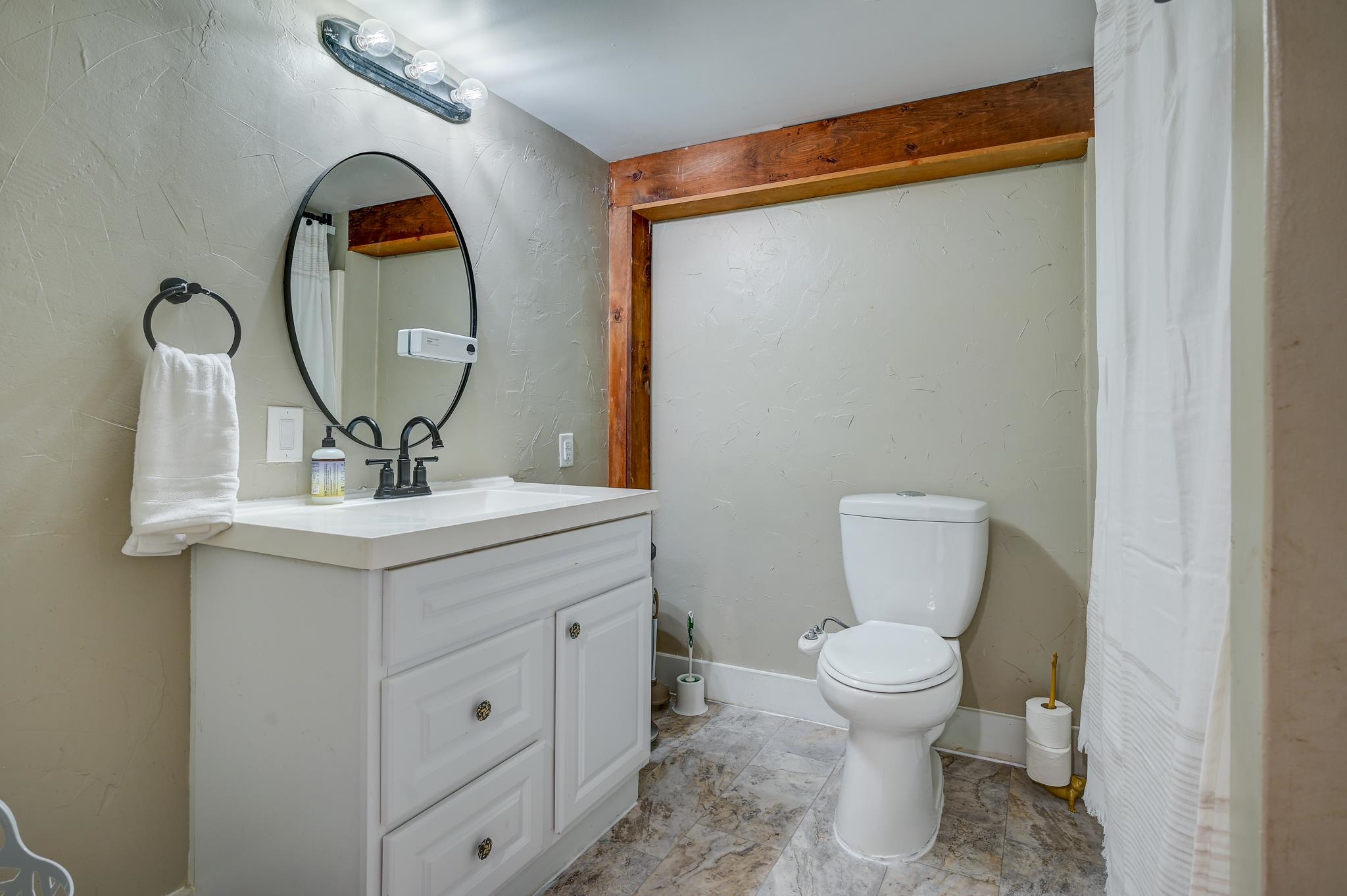 Bathroom with a white vanity, oval mirror, toilet, and exposed wood beam.