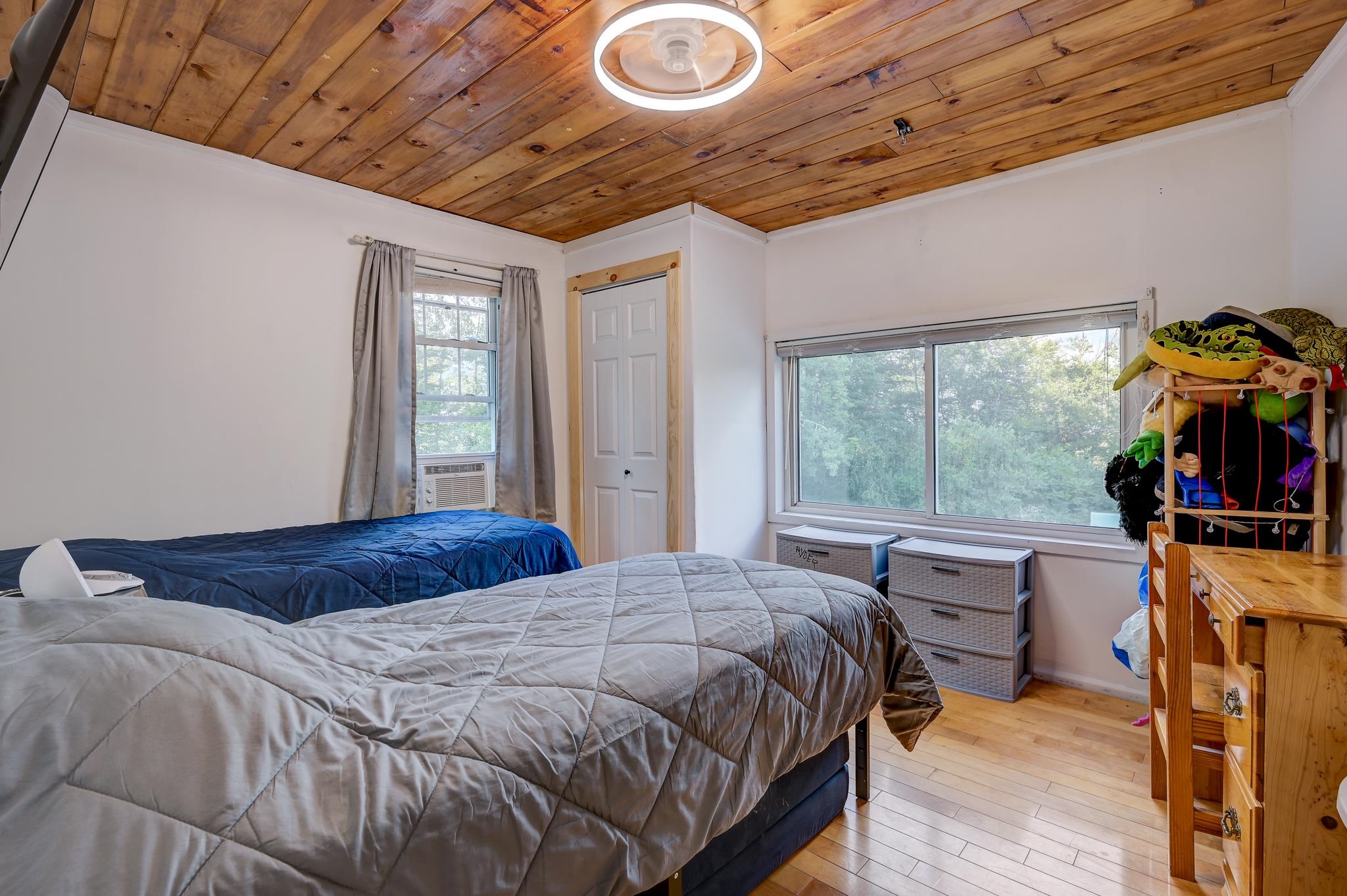 Bedroom with two beds, a wood-plank ceiling, and large windows facing trees.