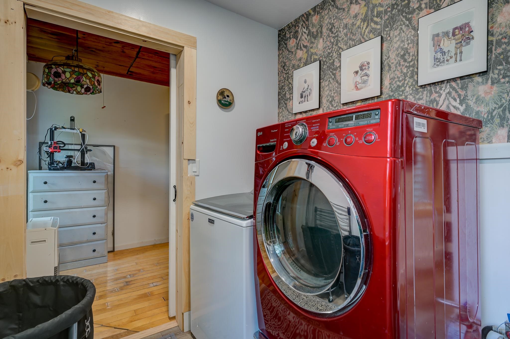 Laundry room with a bright red front-loading washer, white chest-style appliance, and floral wallpaper.