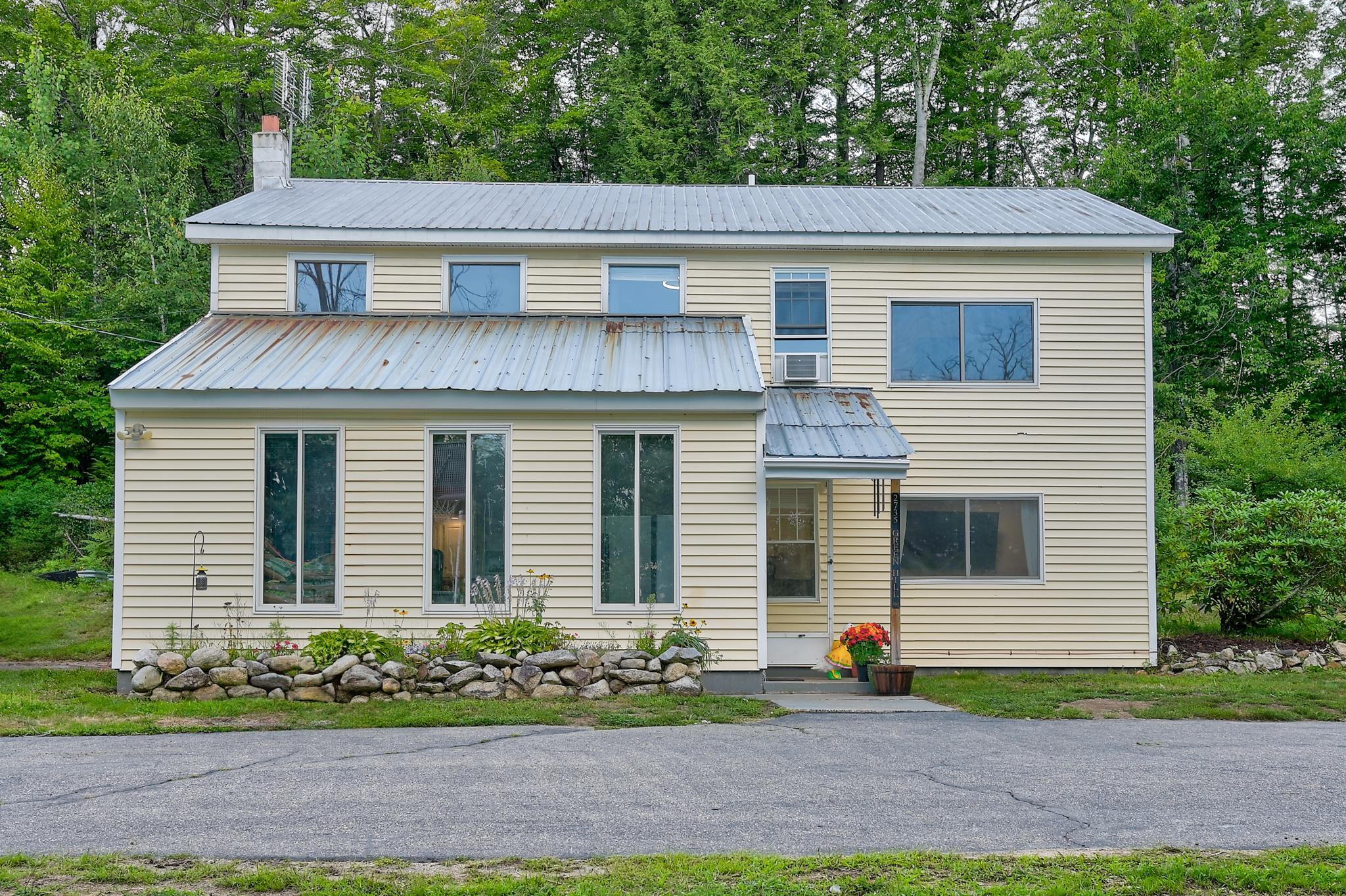 Two-story beige house with a metal roof, stone garden border, and trees behind it.