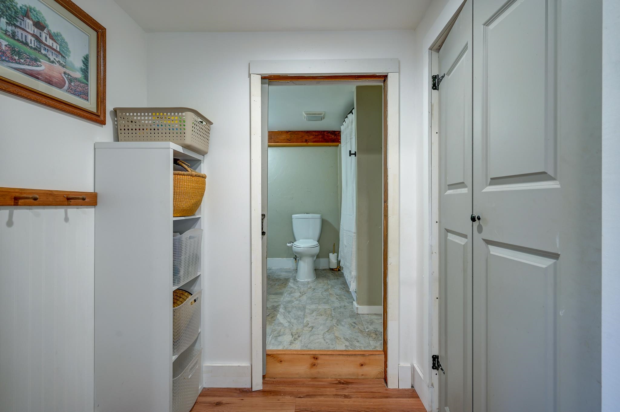 Bathroom with a toilet and shower curtain, viewed past hallway shelving and baskets.
