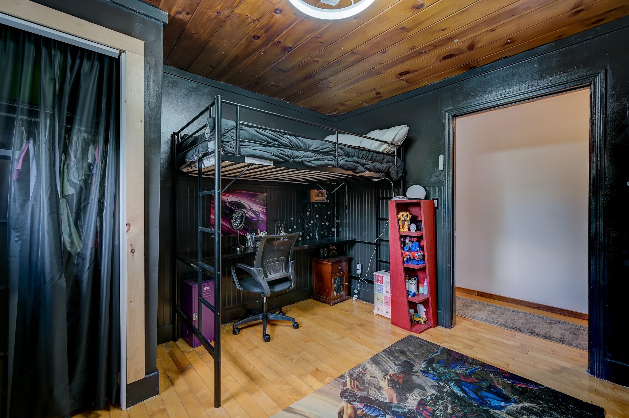 Bedroom with a loft bed over a desk, dark paneled walls, wood ceiling, and red toy shelf.