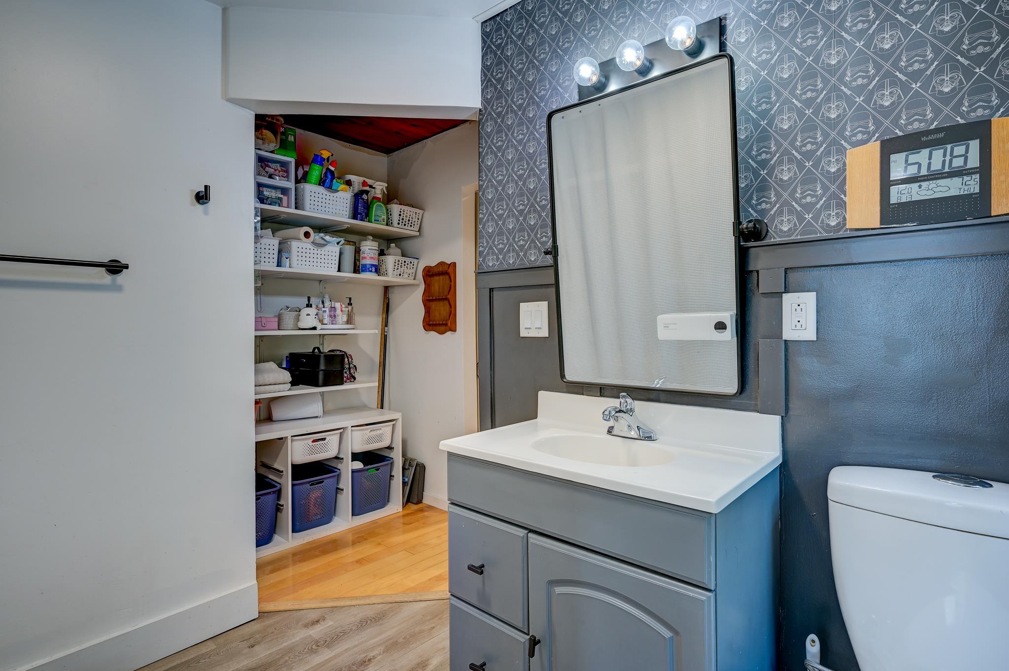 Bathroom with gray vanity, patterned wallpaper, and an open closet filled with organized supplies.