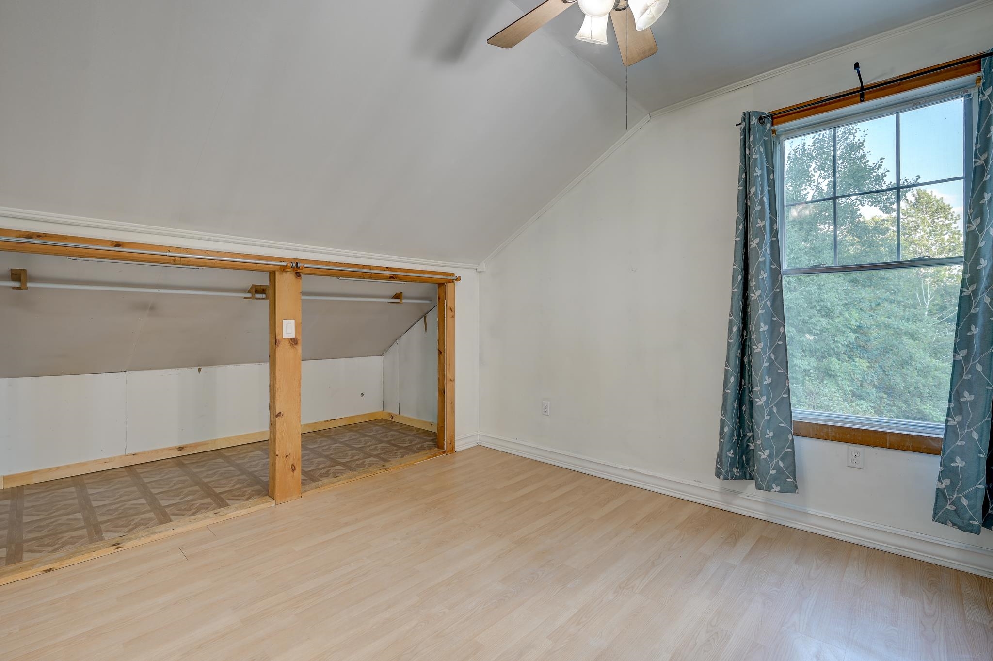 Unfurnished upper-level room with a sloped ceiling, open storage alcove, and large window overlooking trees.