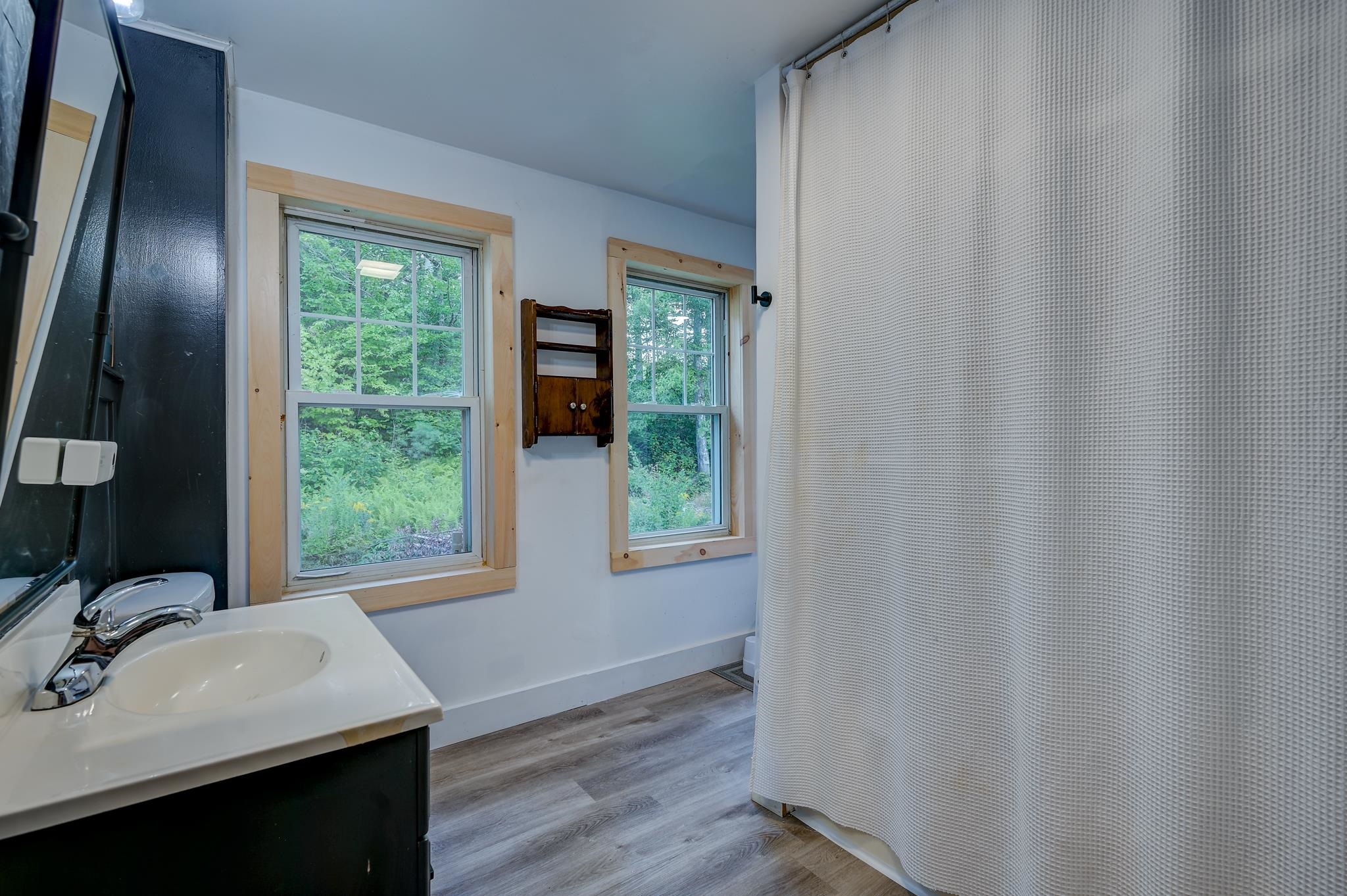 Bathroom with a white sink, wood-trimmed windows overlooking trees, and a textured shower curtain.