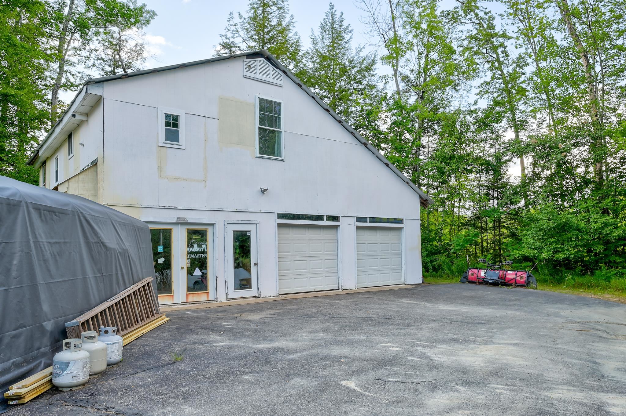White barn-style building with two garage doors, unfinished facade, and paved driveway among trees.