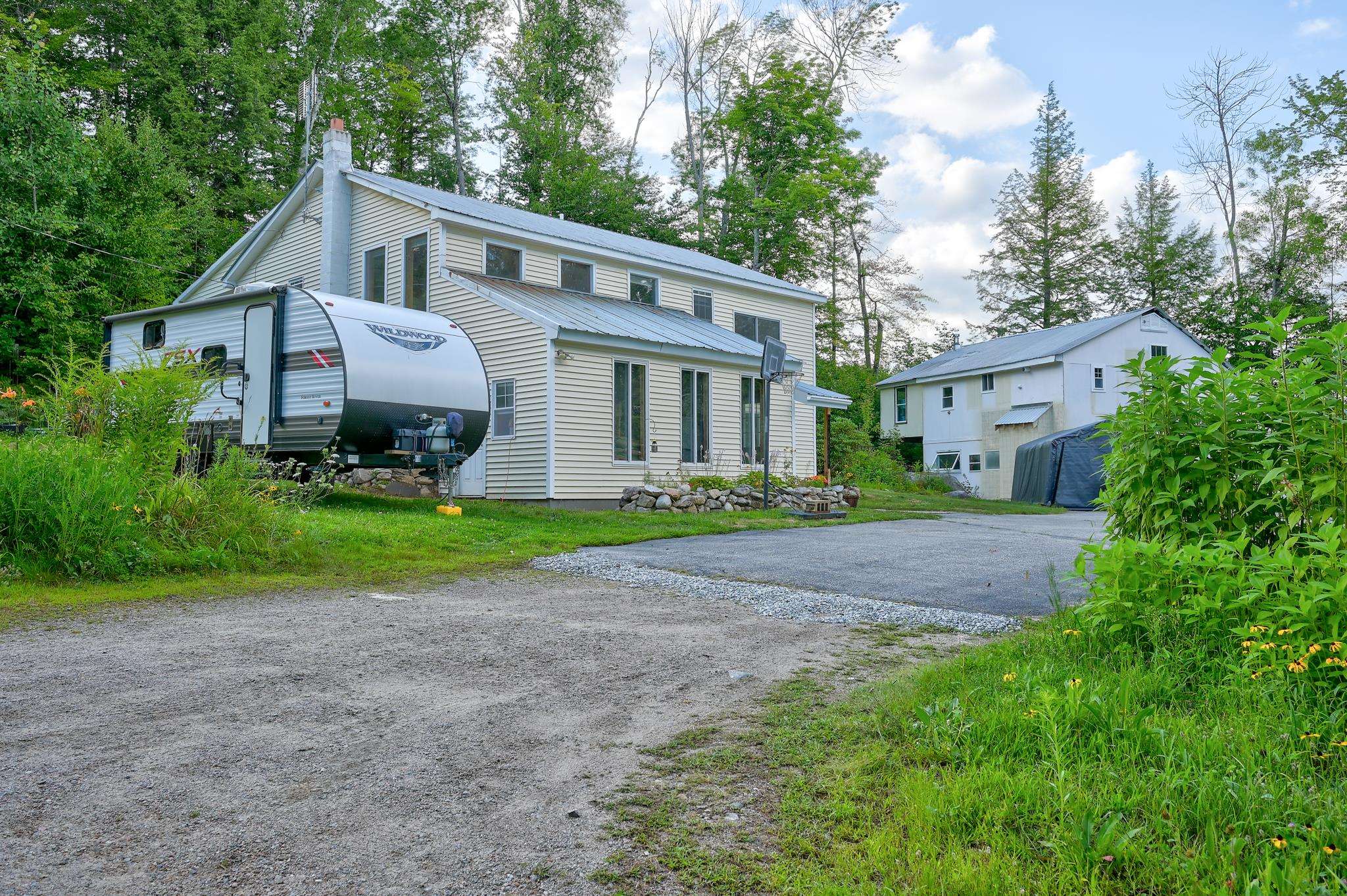 Beige two-story house beside a travel trailer, with a white outbuilding and gravel driveway.