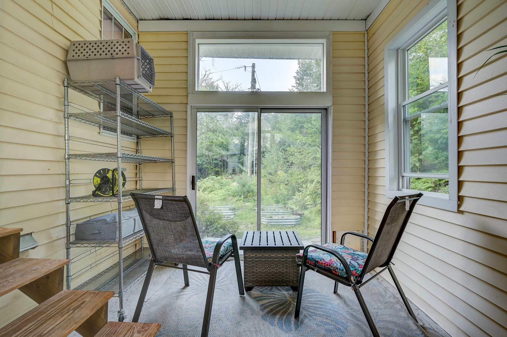 Enclosed porch with two cushioned chairs and a small table facing sliding glass doors and greenery.