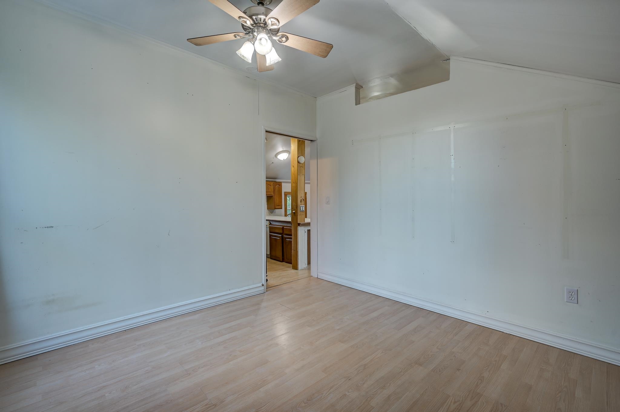 Empty room with pale wood flooring and a ceiling fan, looking through a doorway into the kitchen.