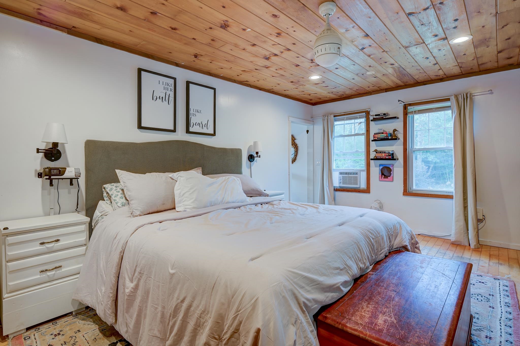 Bedroom with a large white bed beneath a warm wood-plank ceiling and two curtained windows.