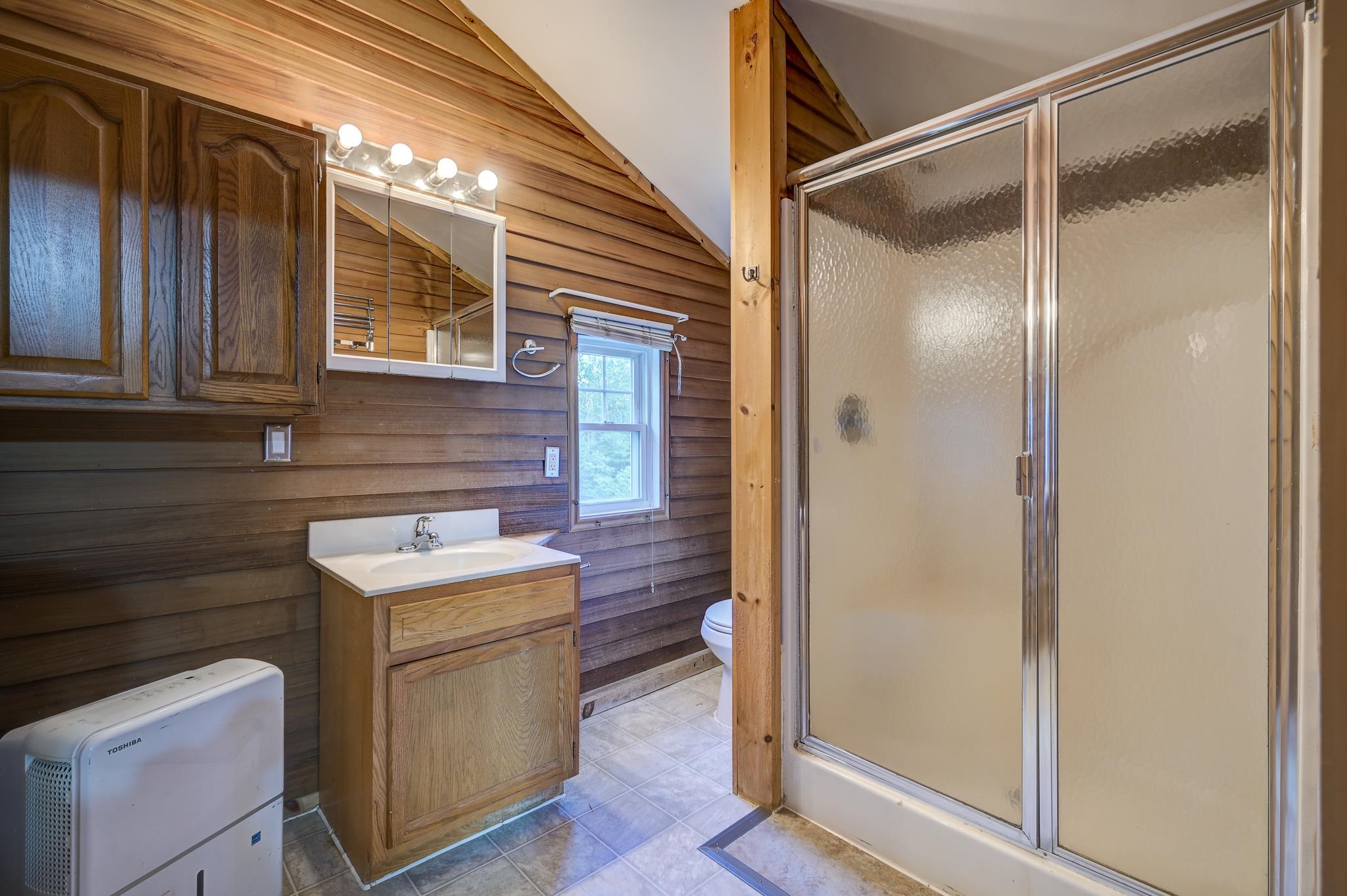 Bathroom with a frosted-glass shower, wood-paneled walls, and a compact vanity beside a window.