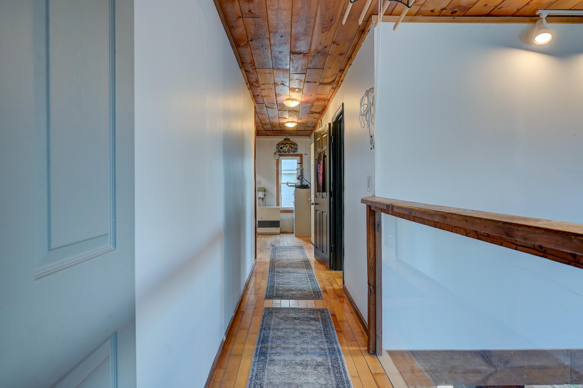 Hallway with a rustic wood ceiling, tile floor, patterned runners, and a wooden stairwell railing.