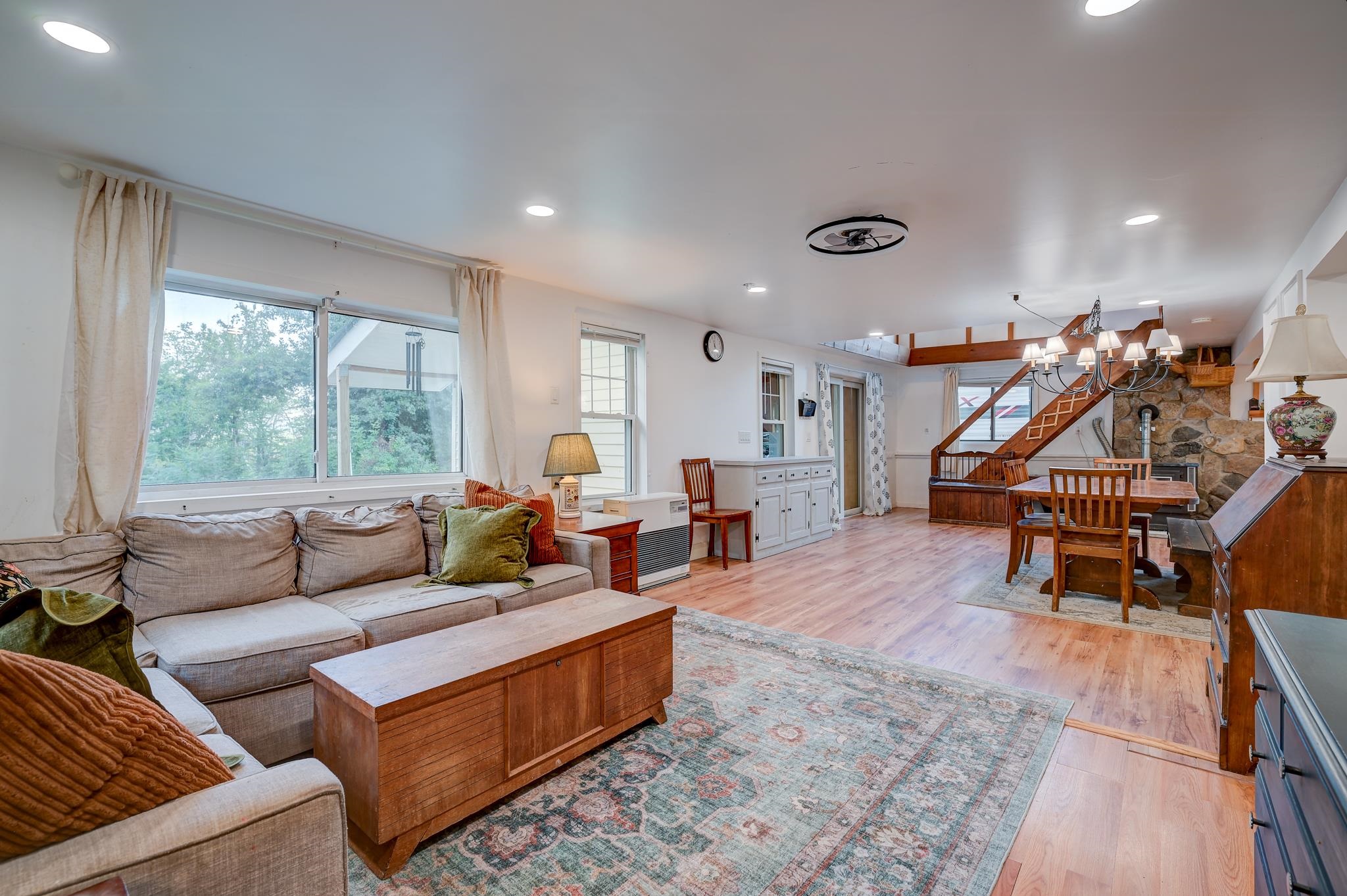 Open-plan living and dining room with a stone fireplace, wooden staircase, and large windows.