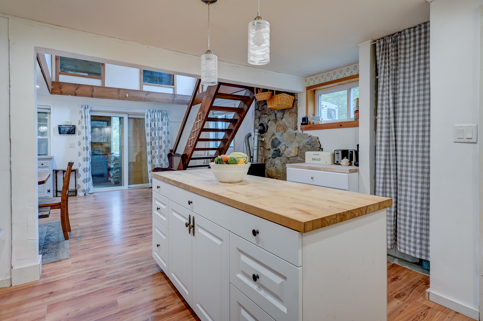 Kitchen with a butcher-block island, loft staircase, and stone wall with a wood stove.