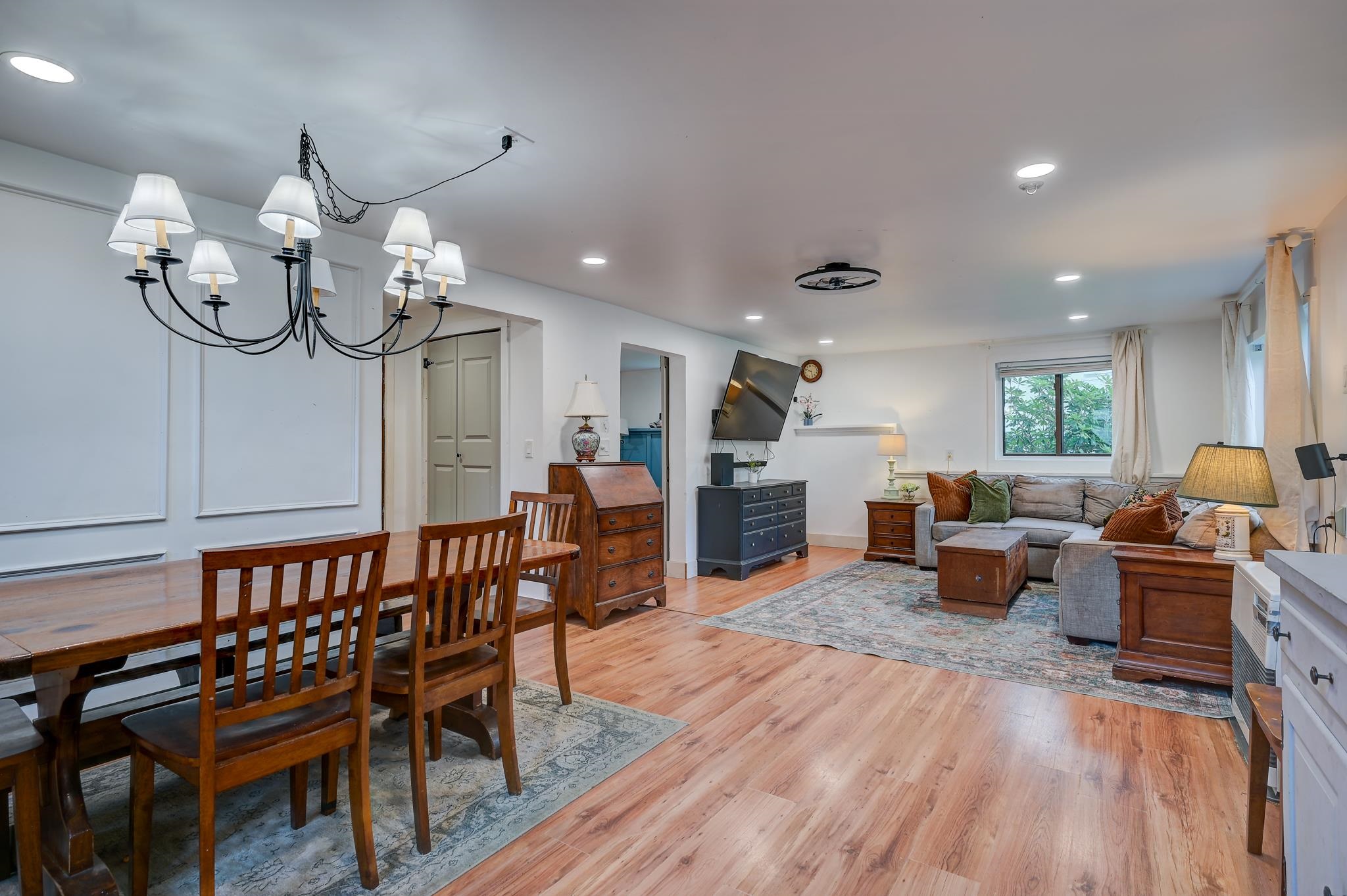 Open-plan living and dining room with wood floors, a chandelier, and a gray sofa by the window.