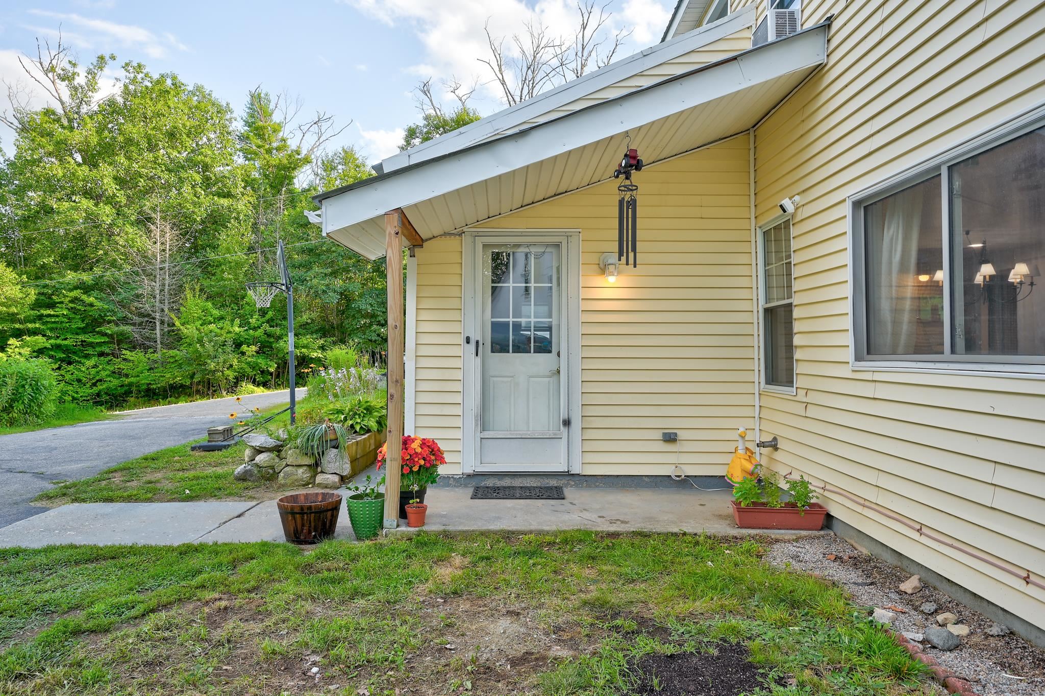 Yellow-sided home’s covered side entrance with flower planters and a basketball hoop nearby.