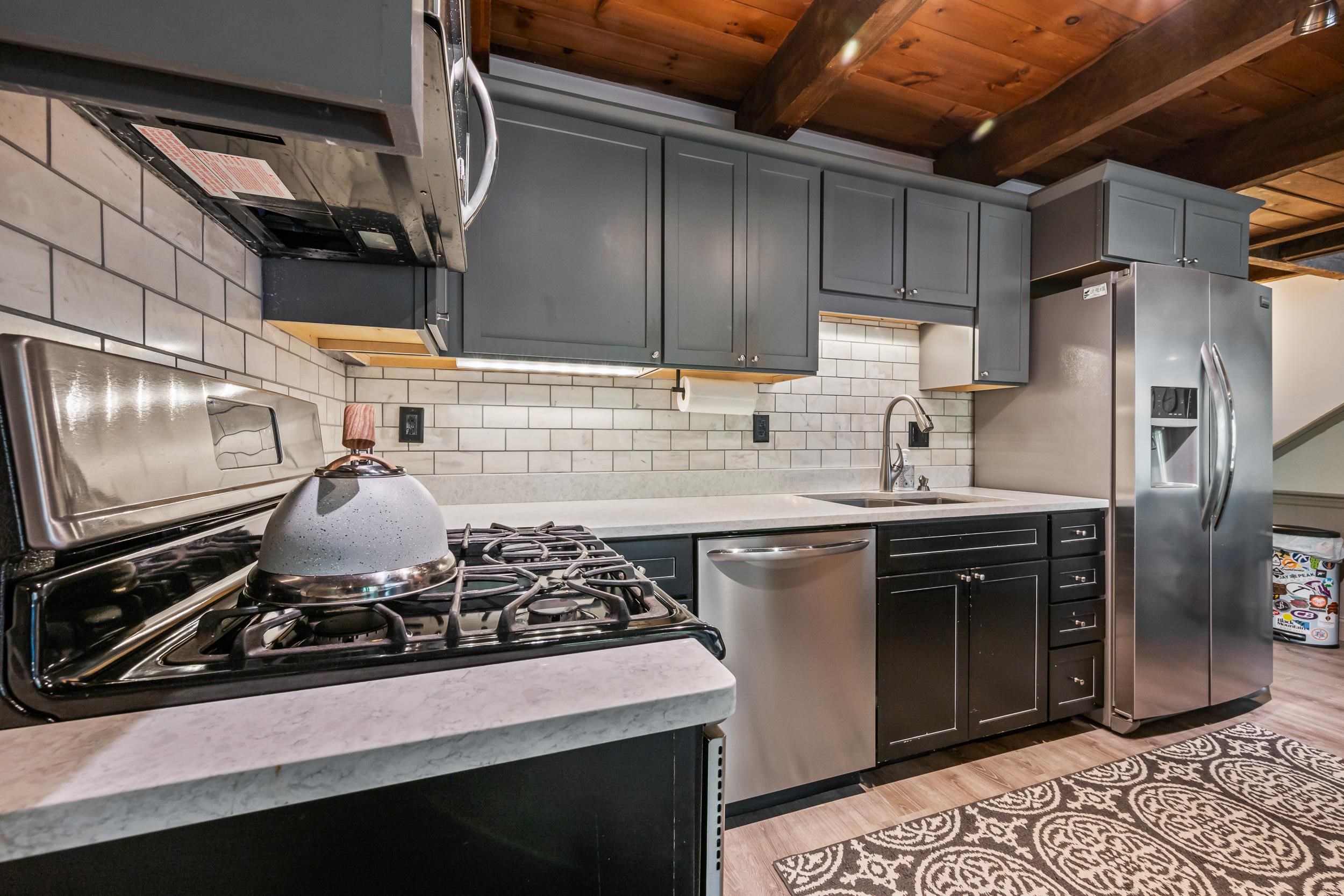 Kitchen with dark gray cabinets, stainless steel appliances, subway tile, and exposed wood ceiling beams.