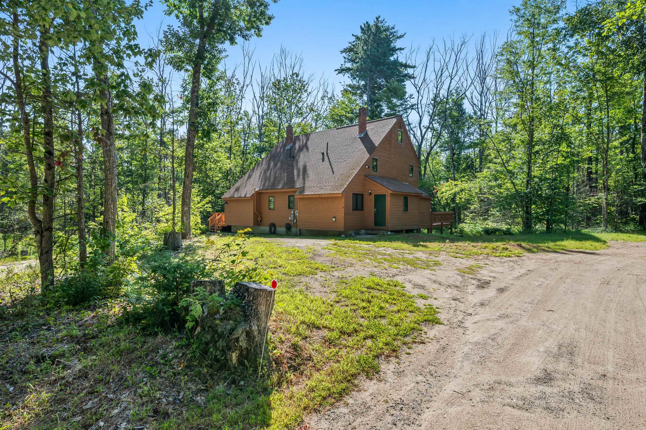 Brown steep-roofed home among trees beside a gravel driveway and small deck.