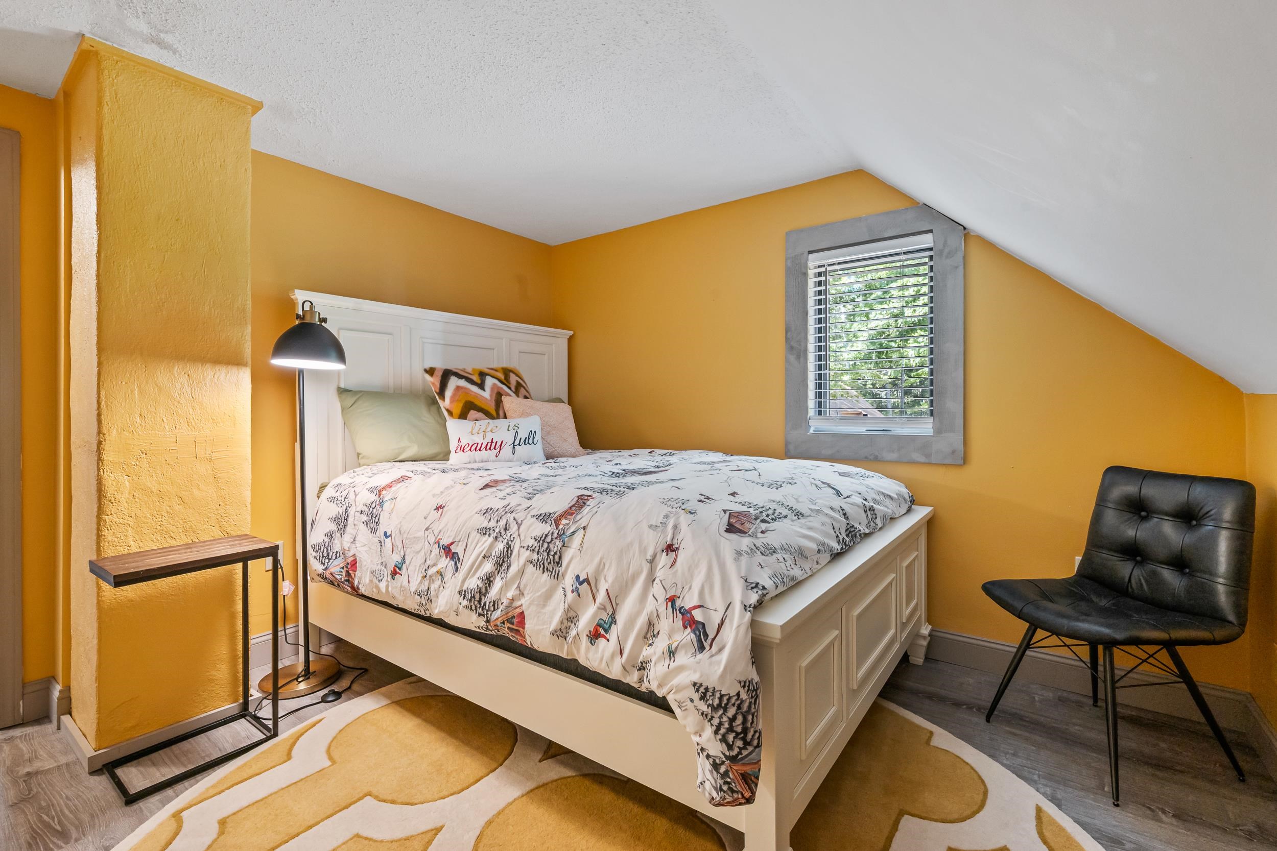 Bedroom with mustard walls, a white bed, patterned bedding, and a black chair beneath a sloped ceiling.